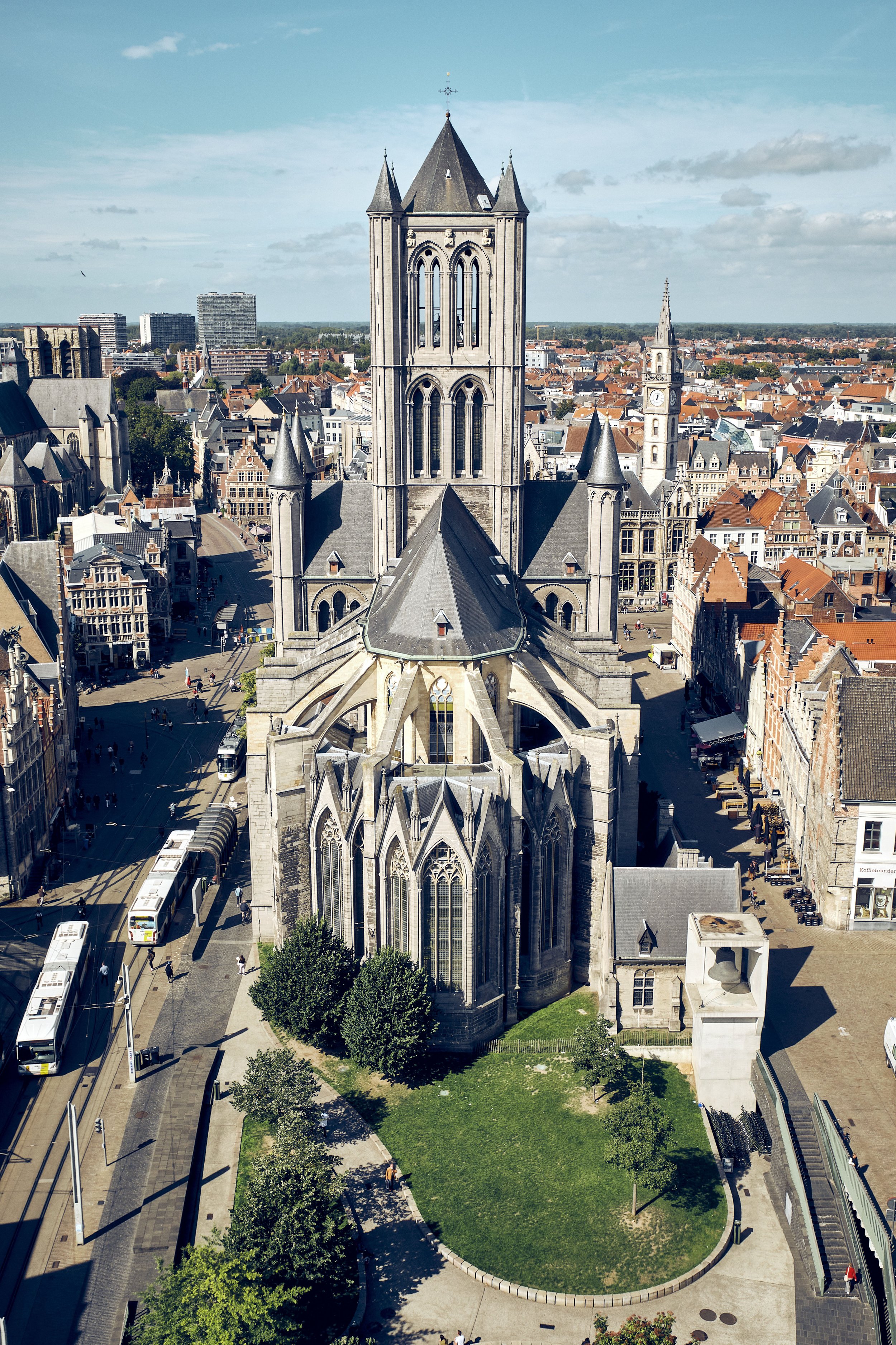 Aerial view of a historic Gothic-style church with a tall central tower, surrounded by a busy city square with people, buses, trees, and surrounding buildings under a partly cloudy sky.