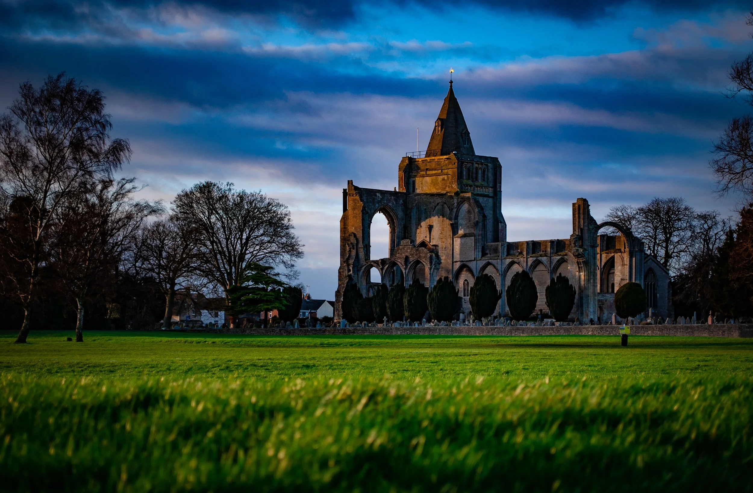 Ruins of an old stone church with a tall steeple, surrounded by trees and a green lawn, under a partly cloudy sky at dusk.
