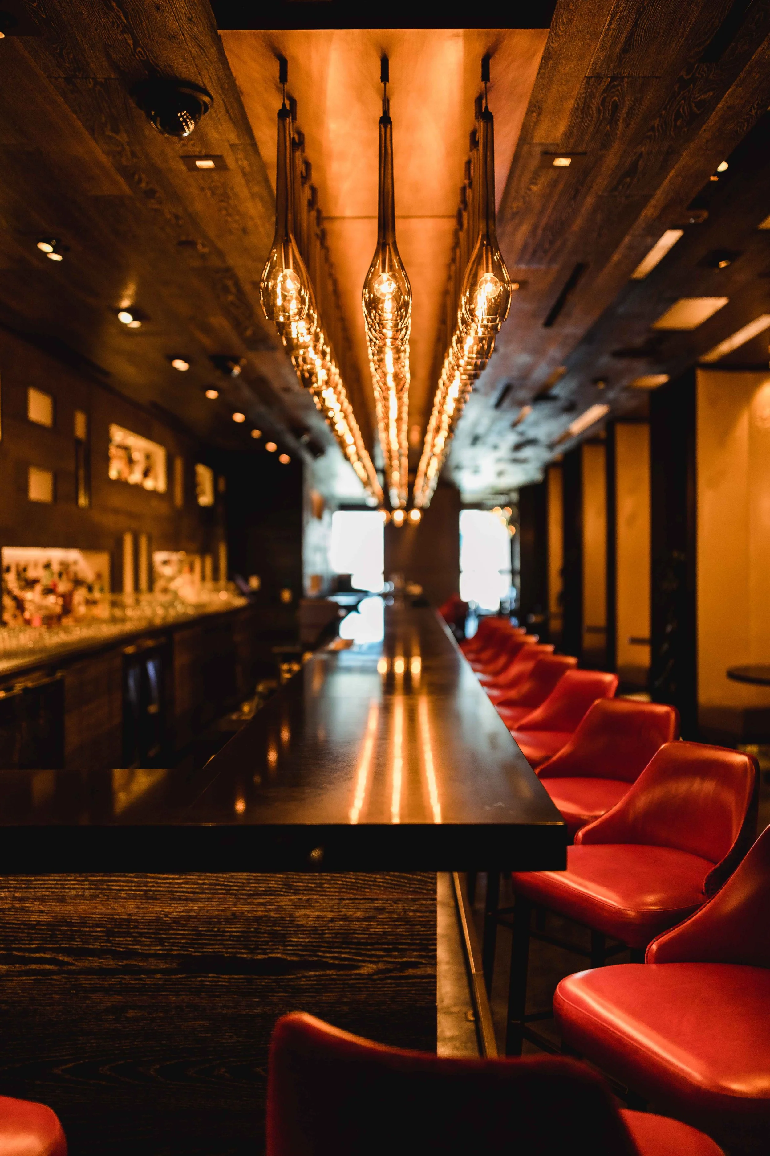 Interior of a dimly lit bar with a long black counter, red bar stools, and a row of hanging pendant lights.