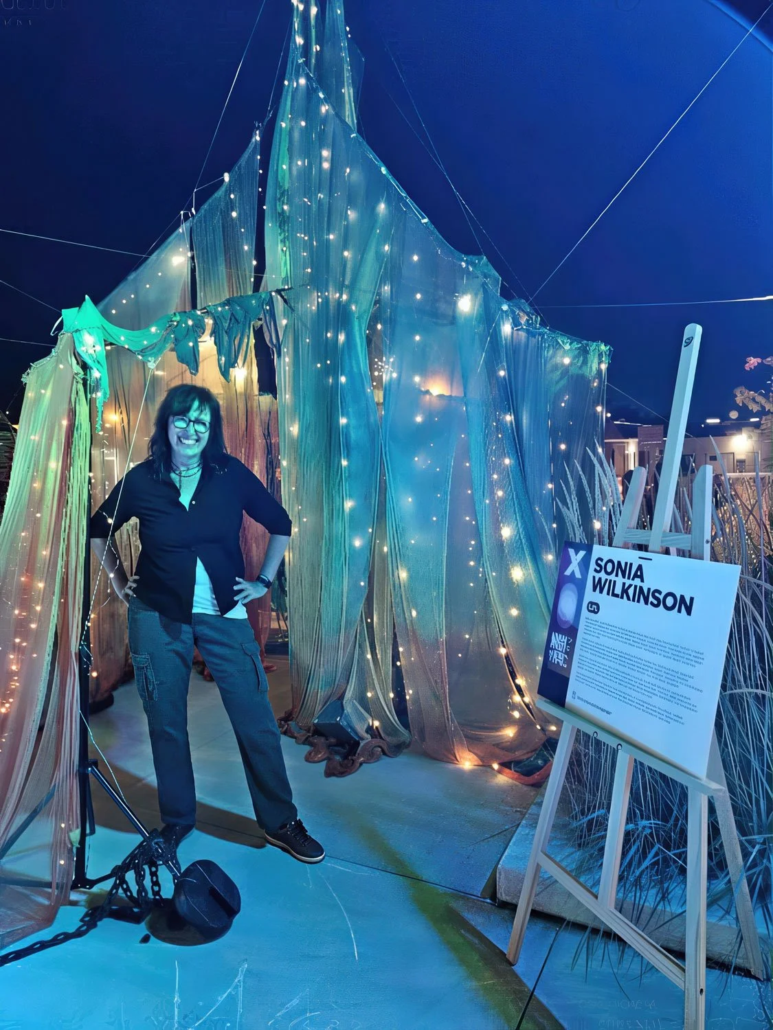 A woman stands inside a glowing art installation made of translucent fabric and fairy lights, next to an informational sign that reads 'Sonia Wilkinson' and features a blue wave graphic.