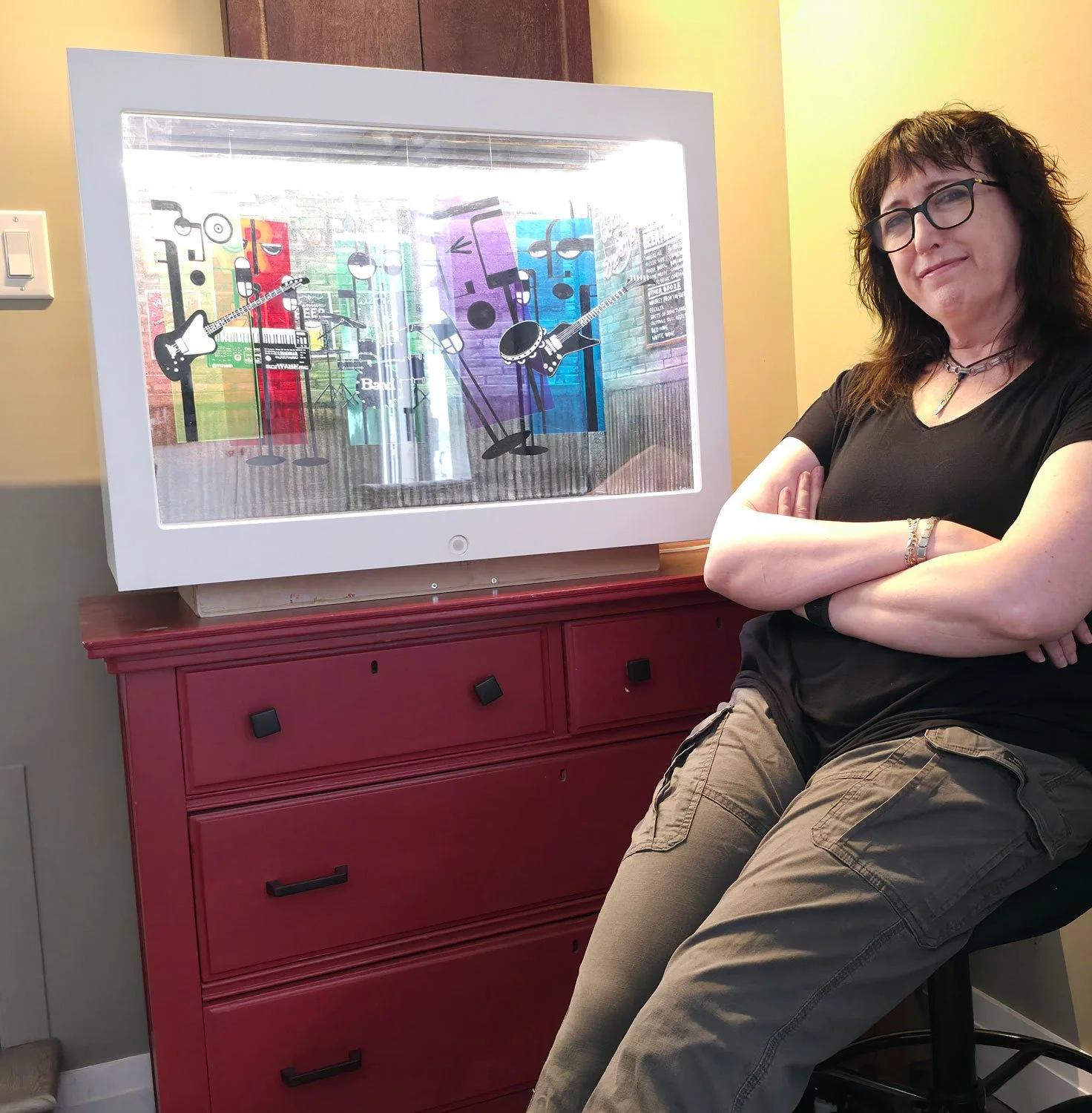 A woman with glasses, dark hair, and layered jewelry sitting with arms crossed next to a television displaying animated music-themed characters.
