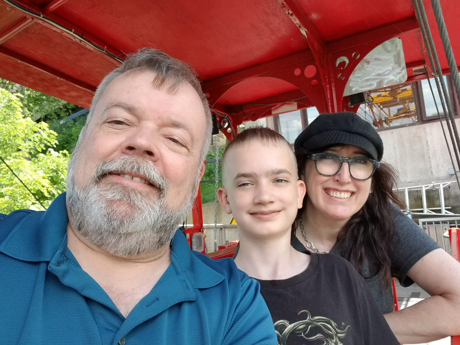 Three people taking a selfie in front of a red amusement park ride, with trees and part of the ride visible in the background.