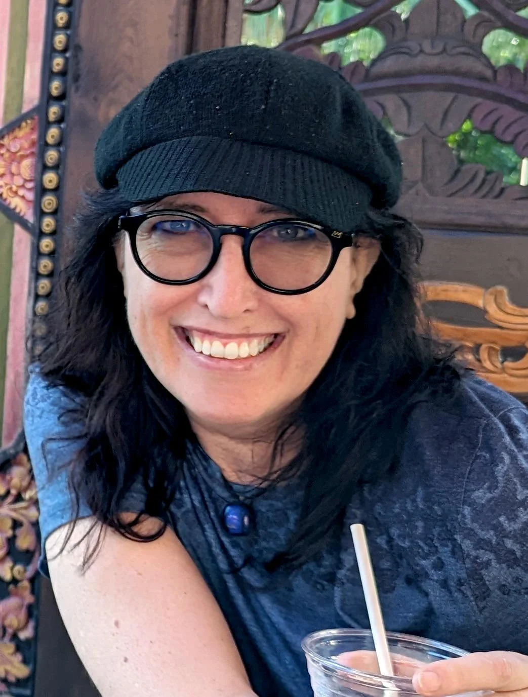 A woman with curly black hair, wearing glasses, a black beret, and a blue shirt, smiling and sitting at a table with a drink in front of her.