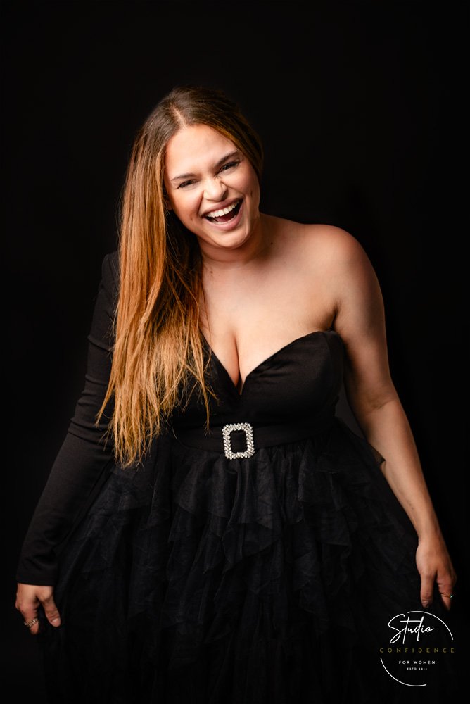 Woman with long hair wearing a strapless black dress with ruffles and a decorative belt, smiling and laughing against a black background.