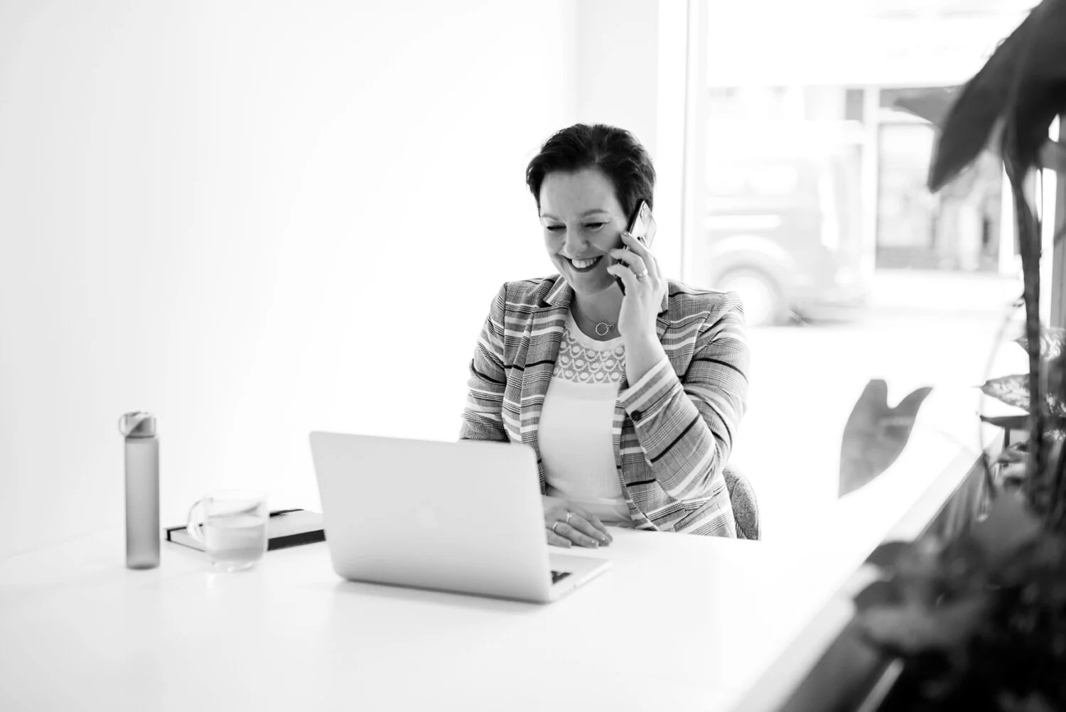 A woman in a blazer smiling on a phone call while sitting at a desk with a laptop, water bottle, and glass of water.