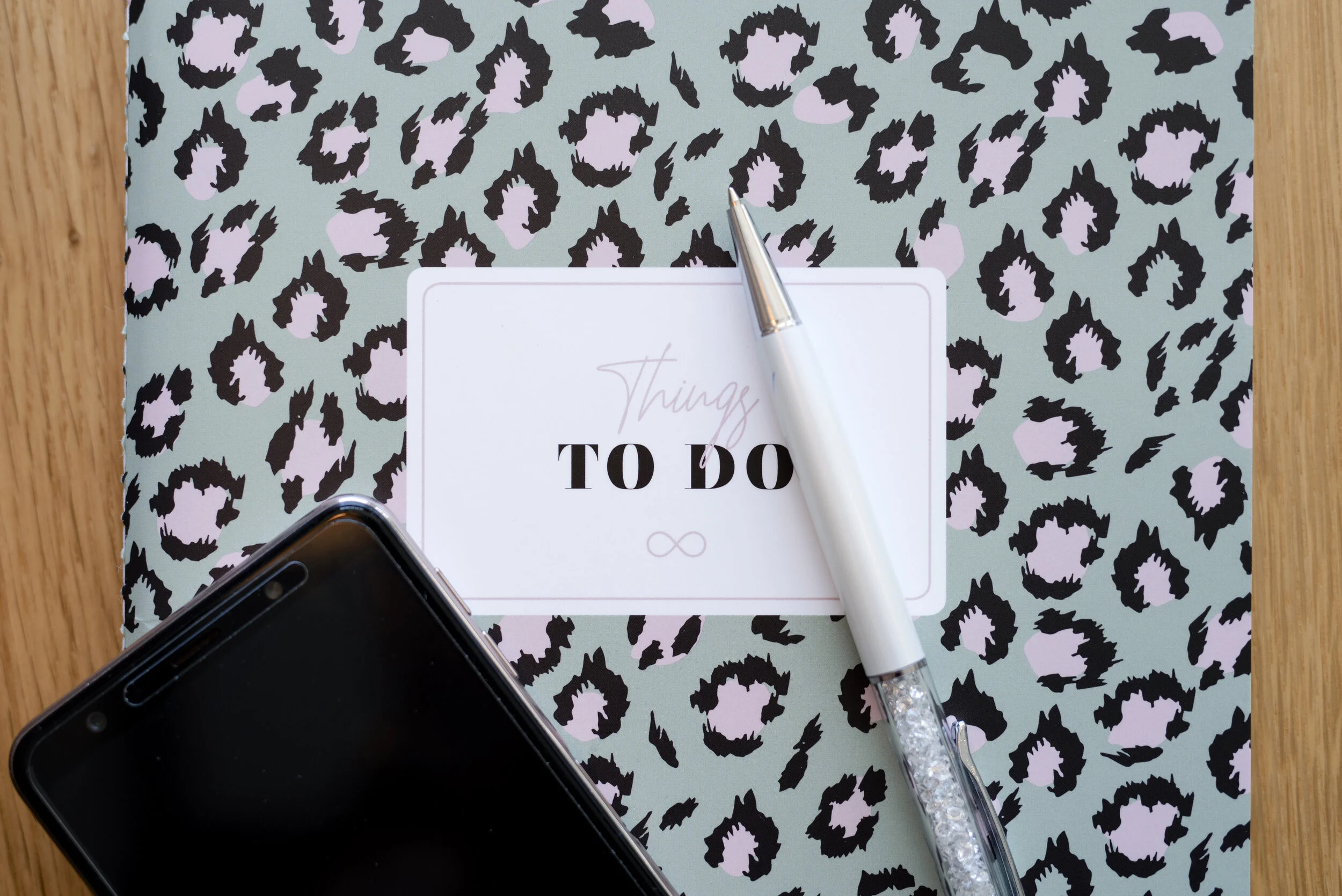 A spiral-bound notebook with a leopard print cover, a white pen with a decorative glitter handle resting on a white and pink 'Things To Do' notepad, and a black smartphone nearby on a wooden surface.