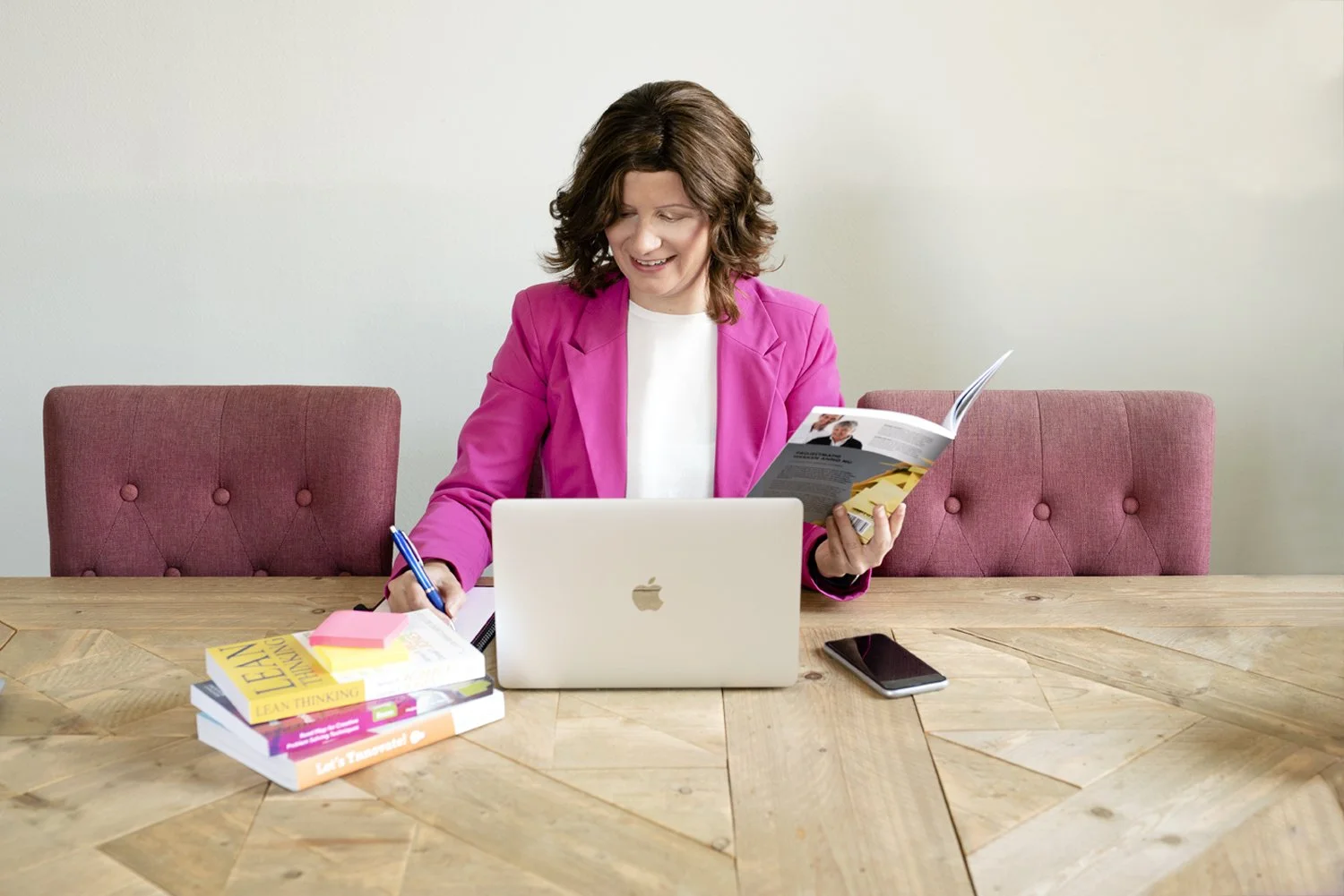 Woman with shoulder-length brown hair in a pink blazer, sitting at a wooden table with a laptop, smartphone, and books, reading a magazine.