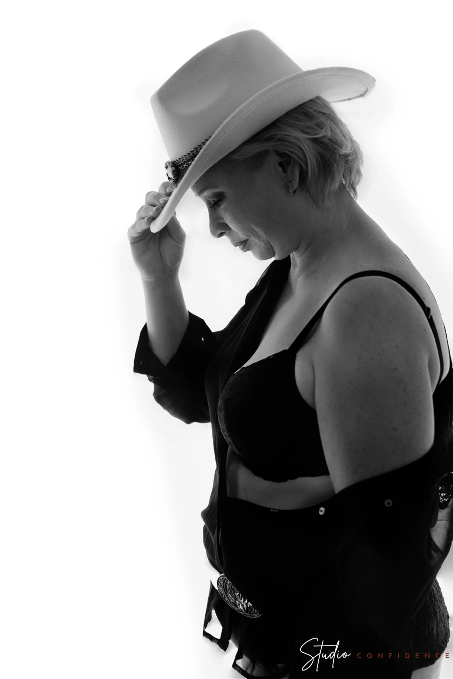Zwart-wit foto van een vrouw met een hoed, zij kijkt naar beneden en houdt de hoed vast. Ze draagt een zwart topje en een rok, met een earring en een leren riem. In de rechter benedenhoek staat de tekst 'Studio Confidence'.