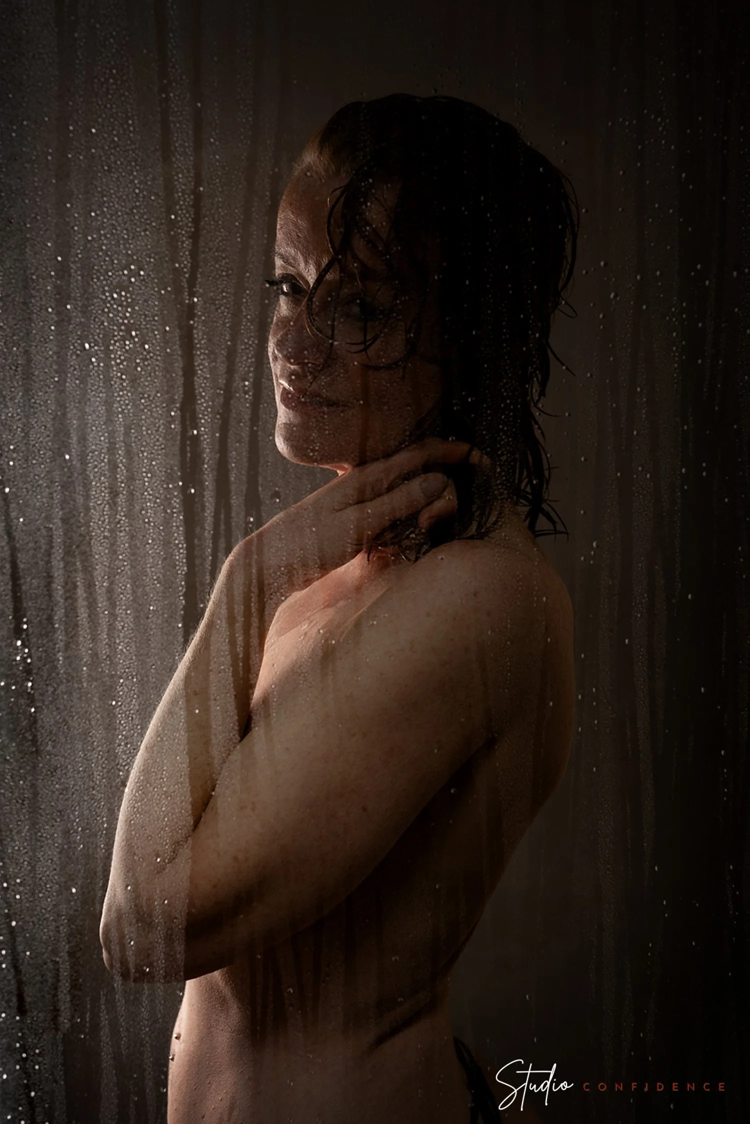 Een vrouw onder de douche met waterdruppels op de ruit en haar huid zichtbaar, met tekst 'Studio Confidence' onderaan.