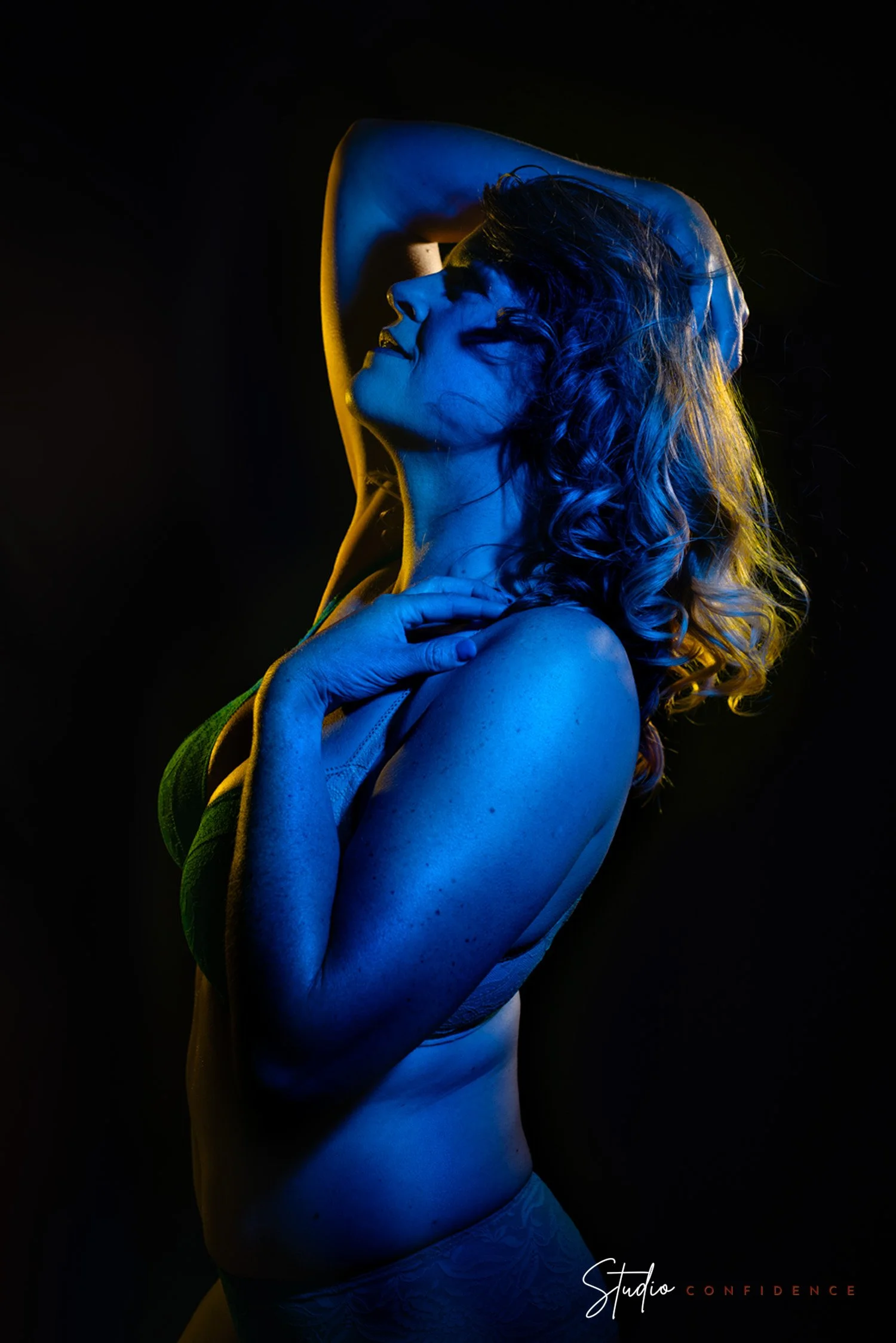 Een vrouw die poseert met een glimlach, haar ogen dicht, met blauwe en gele verlichte belichting, achtergrond is zwart, tekst onderaan leest 'Studio Confidence'