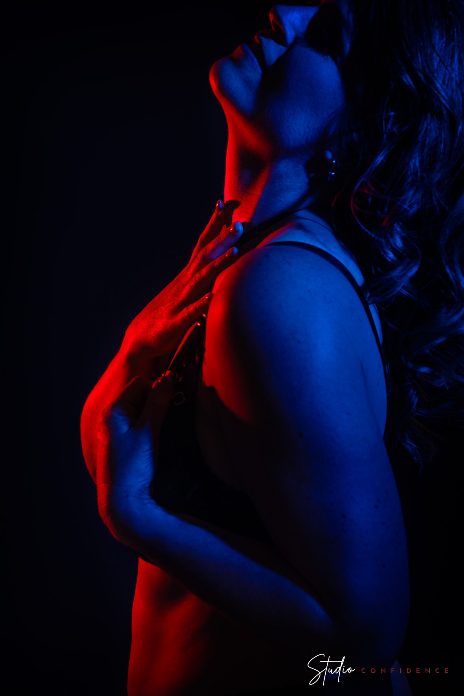 kunstzinnige foto van een vrouw met gesloten ogen, haar gezicht omhoog gericht, verlicht in rood en blauw licht, met de tekst 'Studio Confidence' onderaan.