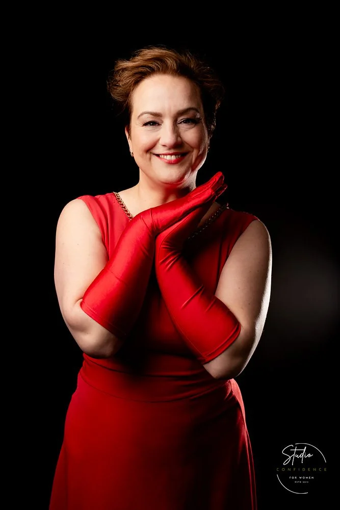 A woman wearing a red dress and matching red gloves, smiling and posing with her hands near her face against a black background.