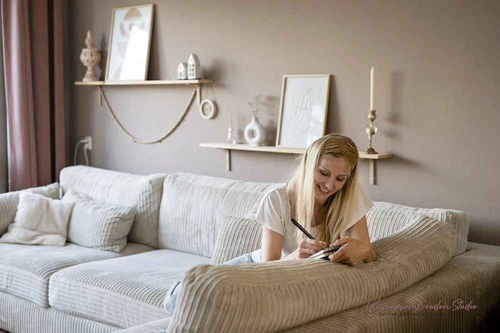 A woman with long blonde hair sitting on a striped beige couch, smiling, writing on a notepad with a black pen in a cozy living room decorated with minimalist art and shelves on the wall.