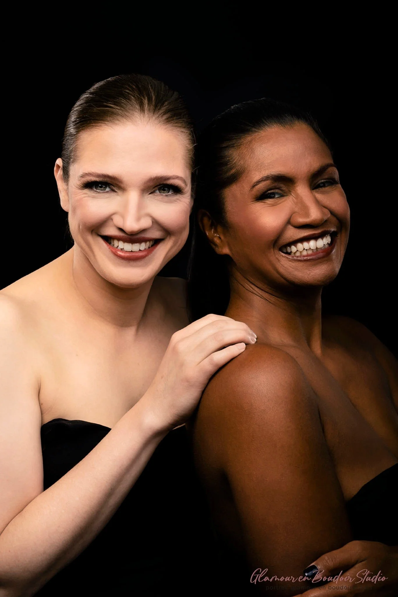 Two women smiling at the camera, one with light skin and the other with dark skin, against a black background.