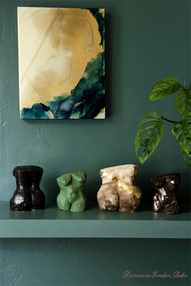 Decorative shelf with four torso-shaped sculptures in black, green, white, and brown, a green potted plant, and abstract watercolor painting on a teal wall.