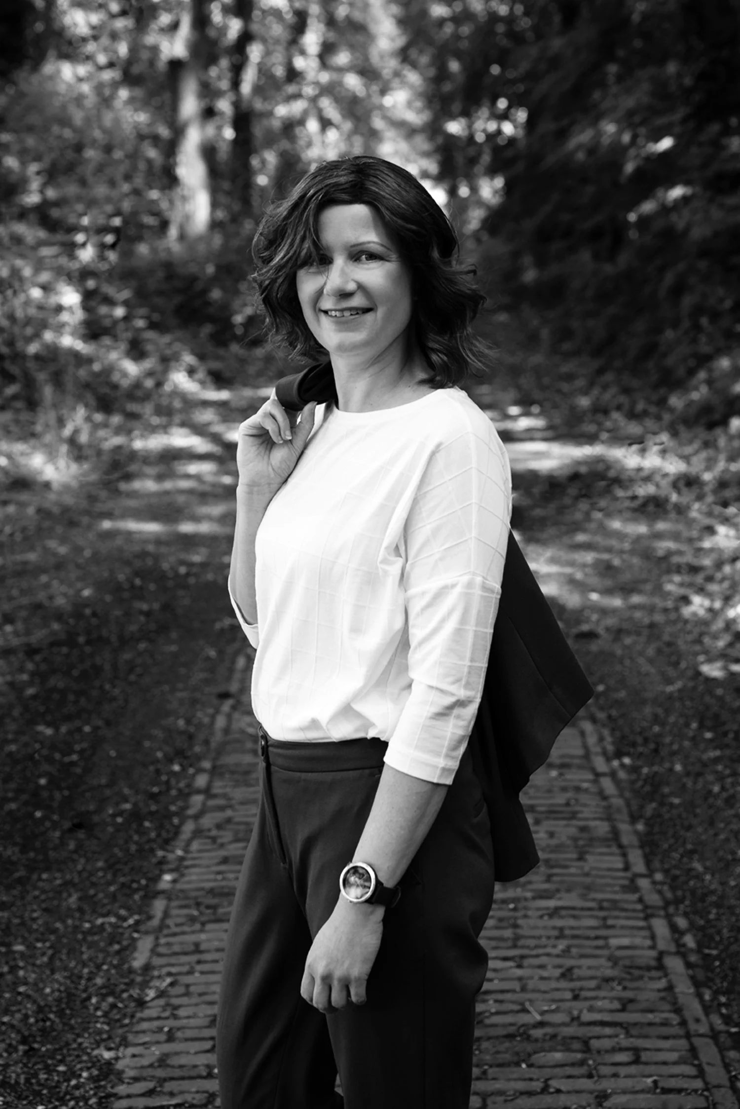 A woman with shoulder-length hair smiling and carrying a backpack over her shoulder, standing on a forest trail.