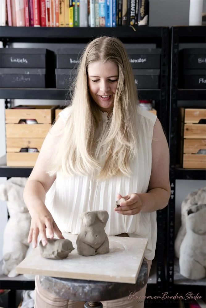 A woman with long blonde hair working on clay sculptures in an art studio, with shelves of books and storage boxes in the background.