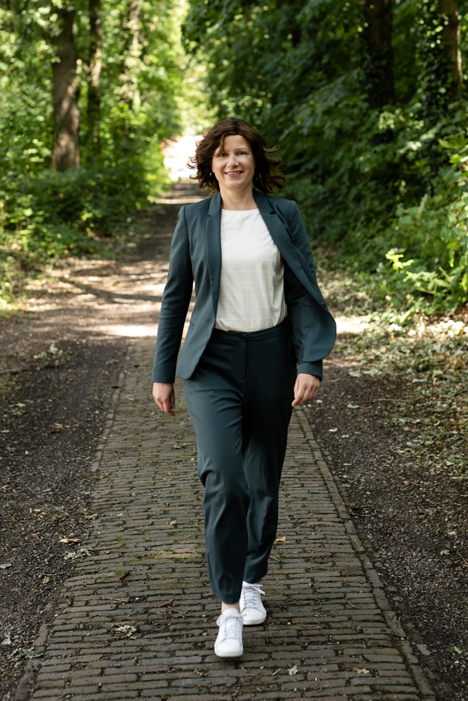 A woman walking on a brick path through a green wooded area, smiling and dressed in business casual attire with a blazer and sneakers.