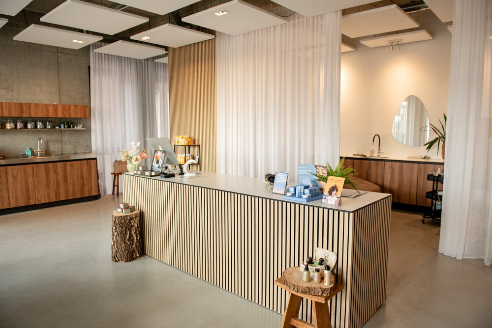 Modern reception desk with wood and white striped design, decorated with skincare products and promotional materials, in a contemporary interior with wooden accents, white curtains, and a small sitting area.