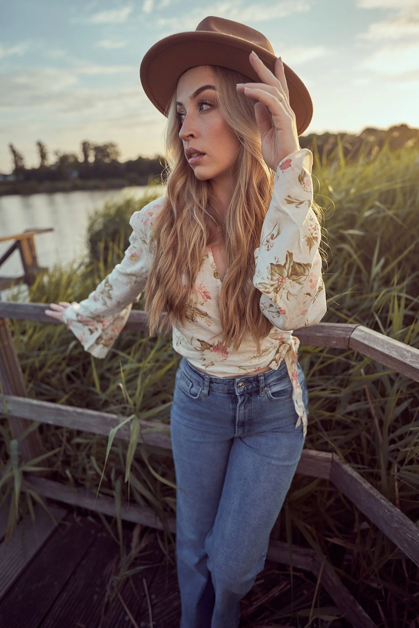 A blonde woman in a studded wide-brim hat, floral blouse, and jeans stands outdoors touching her hat while looking off-camera.