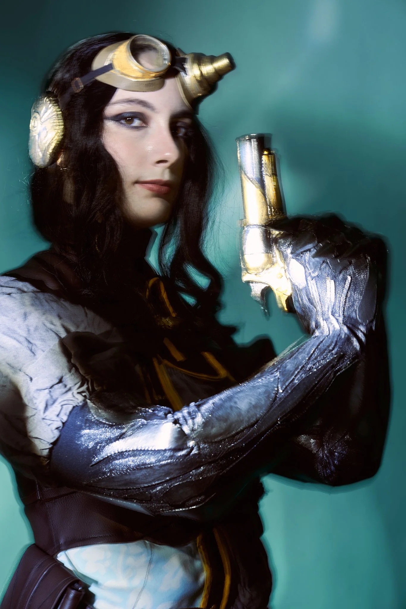 Lady Mechanika cosplay portrait — woman in brass aviator goggles with a silver mechanical arm holding a steampunk pistol, photographed with light-painting technique on a teal backdrop. Editorial photography by Simon Rogghe.