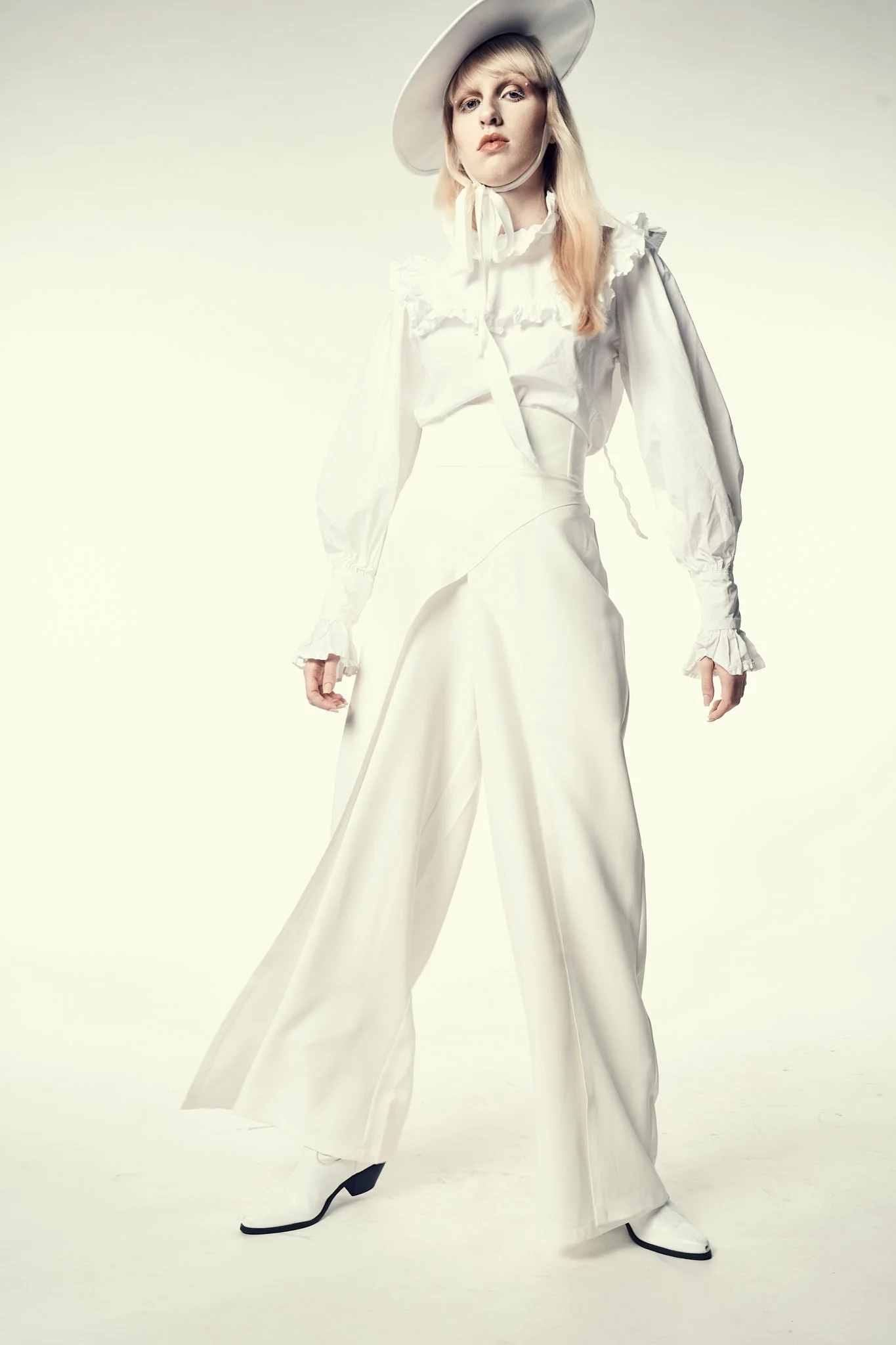 Full-length Western fashion portrait of a blonde model dressed in an all-white ruffled blouse, wide-leg trousers, white boots, and a white hat.⁠