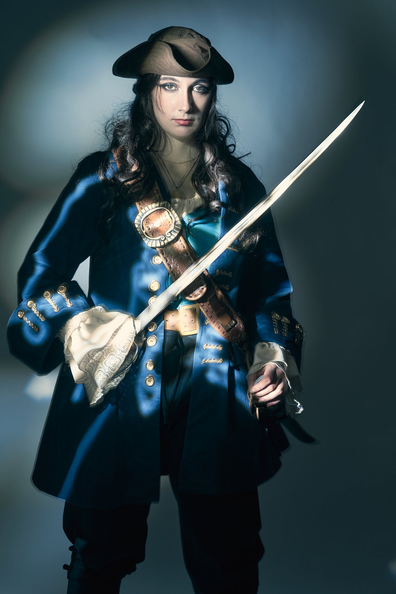 Painterly pirate cosplay portrait — woman in a blue Renaissance-era pirate coat with gold buttons and tricorn hat, holding a sword, photographed with light-painting technique. Editorial cosplay photography by Simon Rogghe.