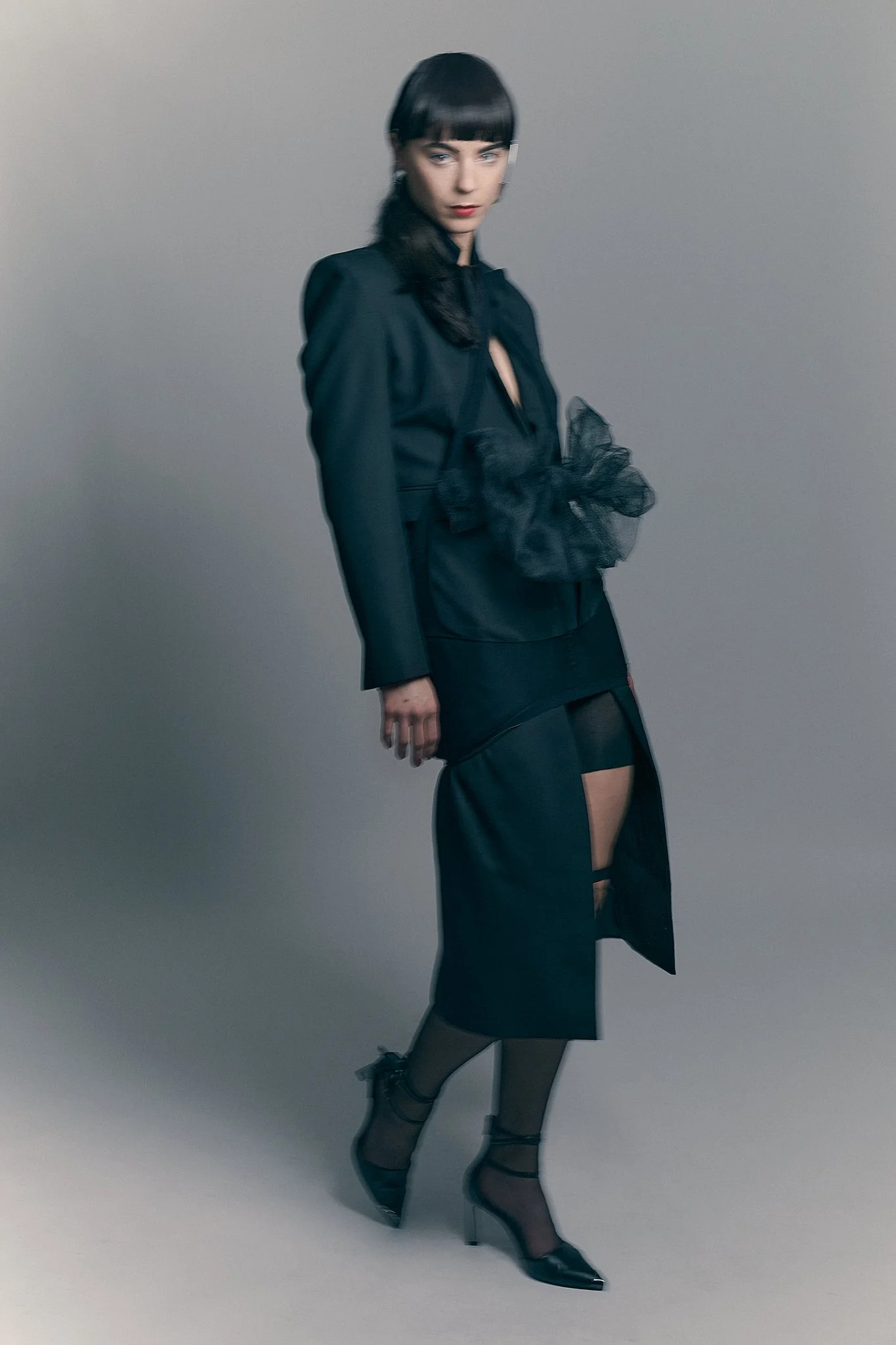 ⁠Full-body editorial portrait of a model with bangs wearing a structured black blazer suit with exaggerated fabric detailing.⁠