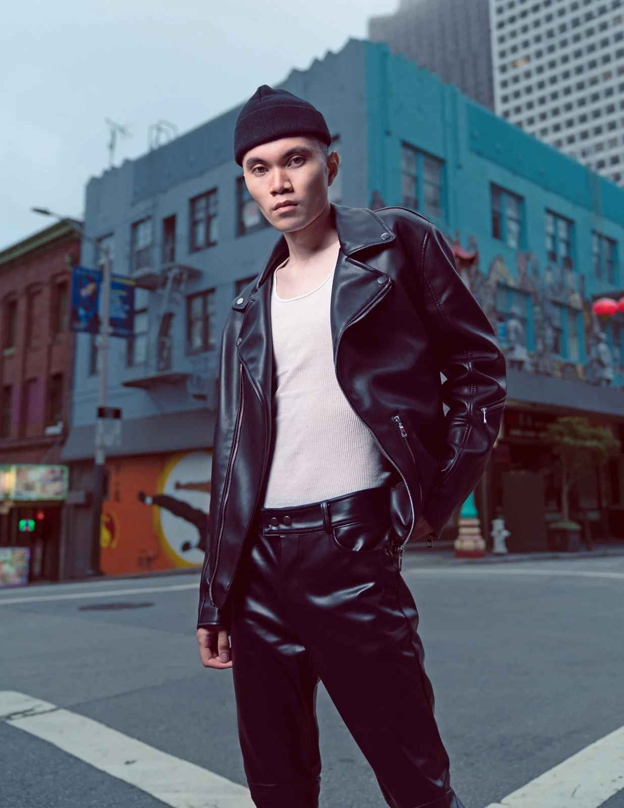 Three-quarter outdoor editorial fashion photograph of a male model posing in a black leather jacket, white tank top, and black trousers on a city street by editorial photographer Simon Rogghe.