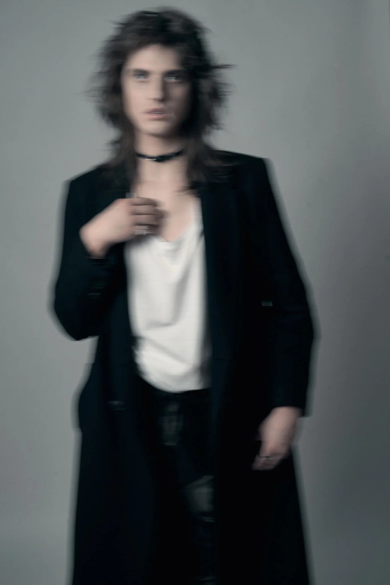Edgy fashion editorial photograph with slight motion blur showing a male model with shag hair wearing an open black coat over a white top and a black choker.⁠
