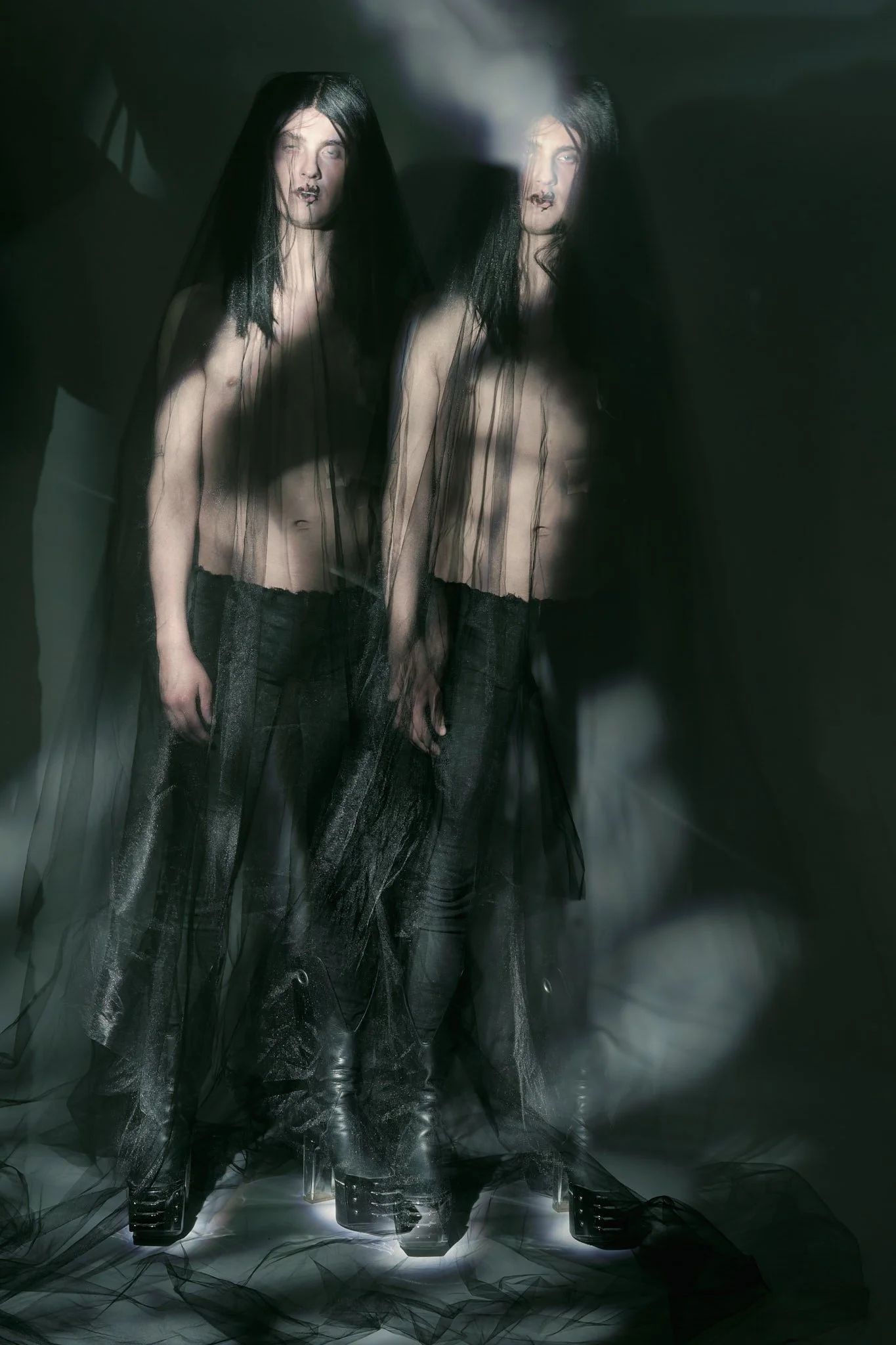 Full-length dark editorial photograph of two shirtless male models draped in sheer black tulle veils and black trousers in a atmospheric studio setting.⁠