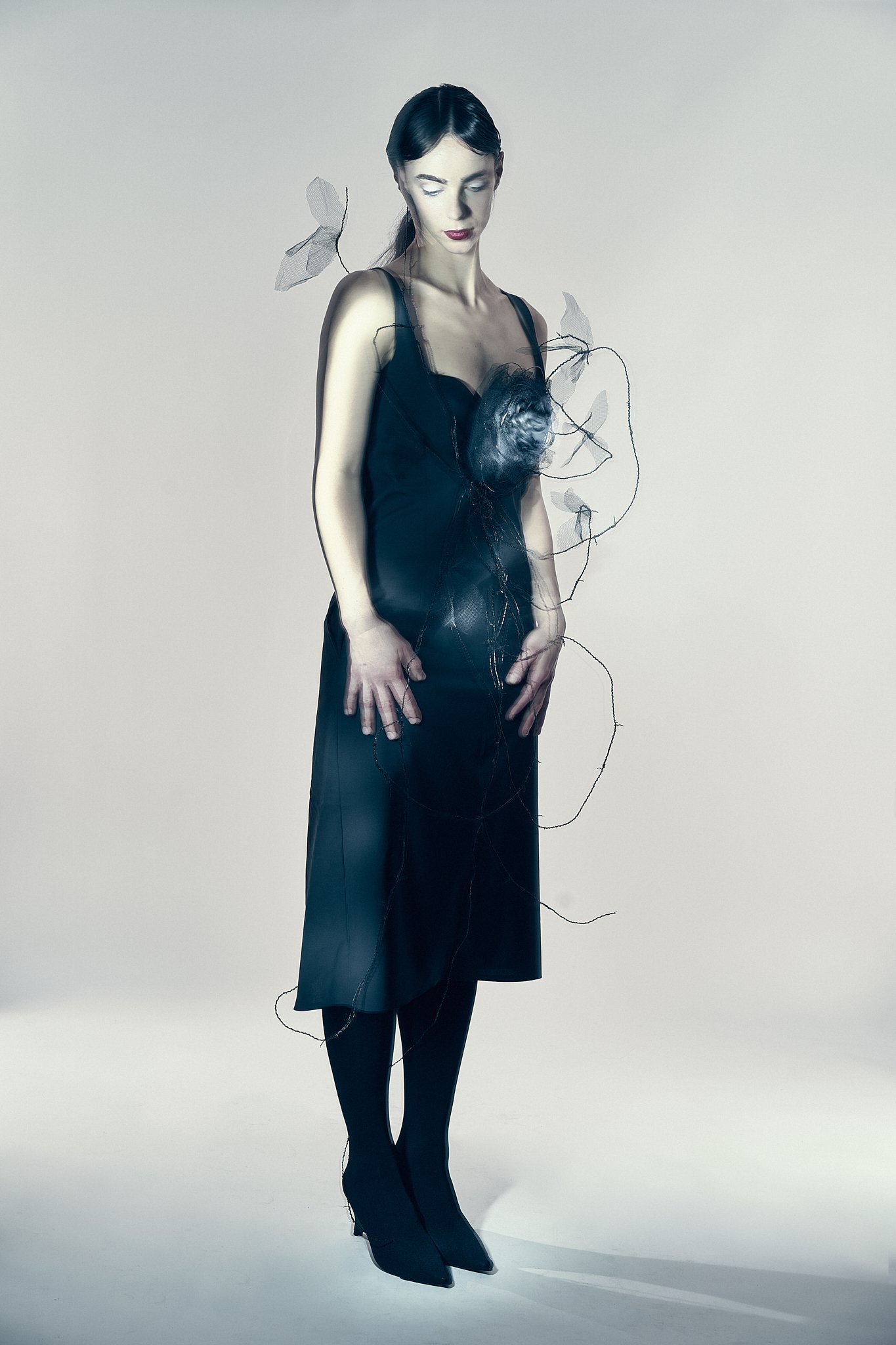 Full-length studio portrait of a model wearing a black mid-length dress with wire botanical elements and dark tights.⁠