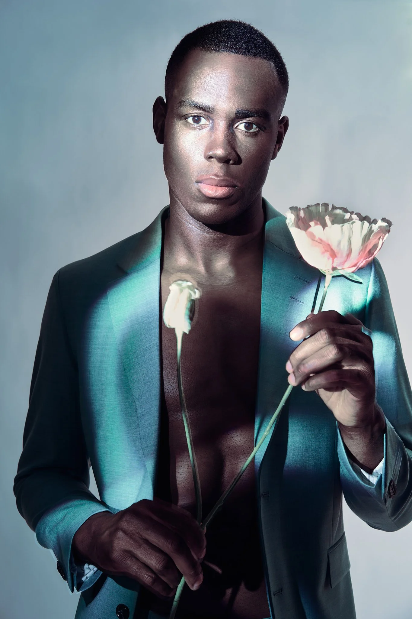 Close-up fashion portrait of a Black male model in a teal blazer, shirtless, holding a single white flower with soft dramatic lighting.⁠