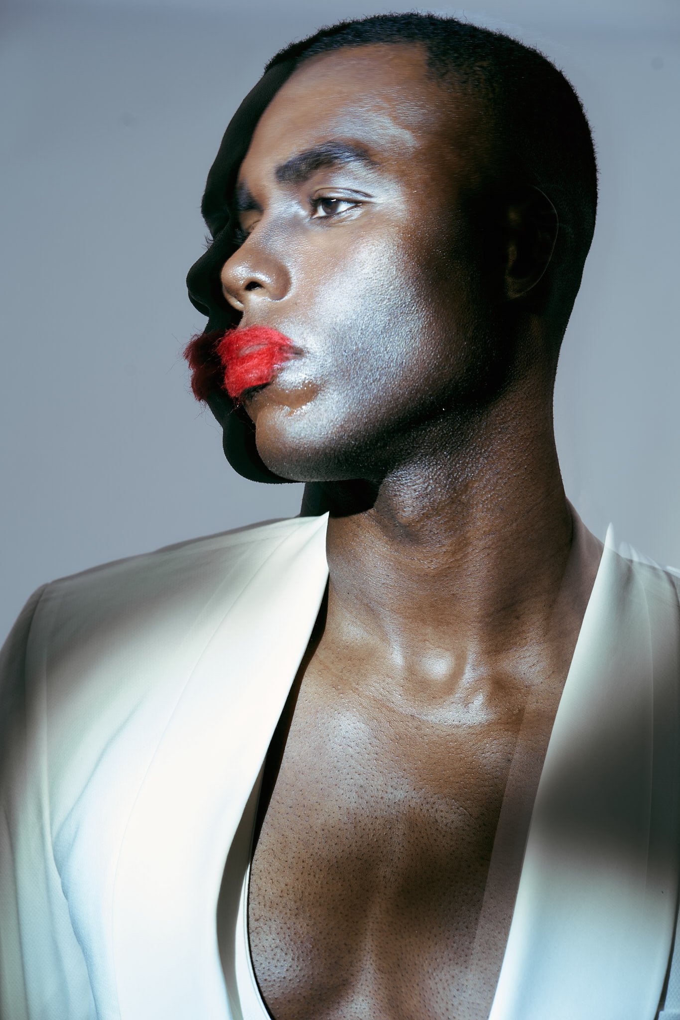 Profile fashion portrait of a Black male model with red fuzzy lips, shirtless in a white blazer, in a soft-focus studio setting.⁠