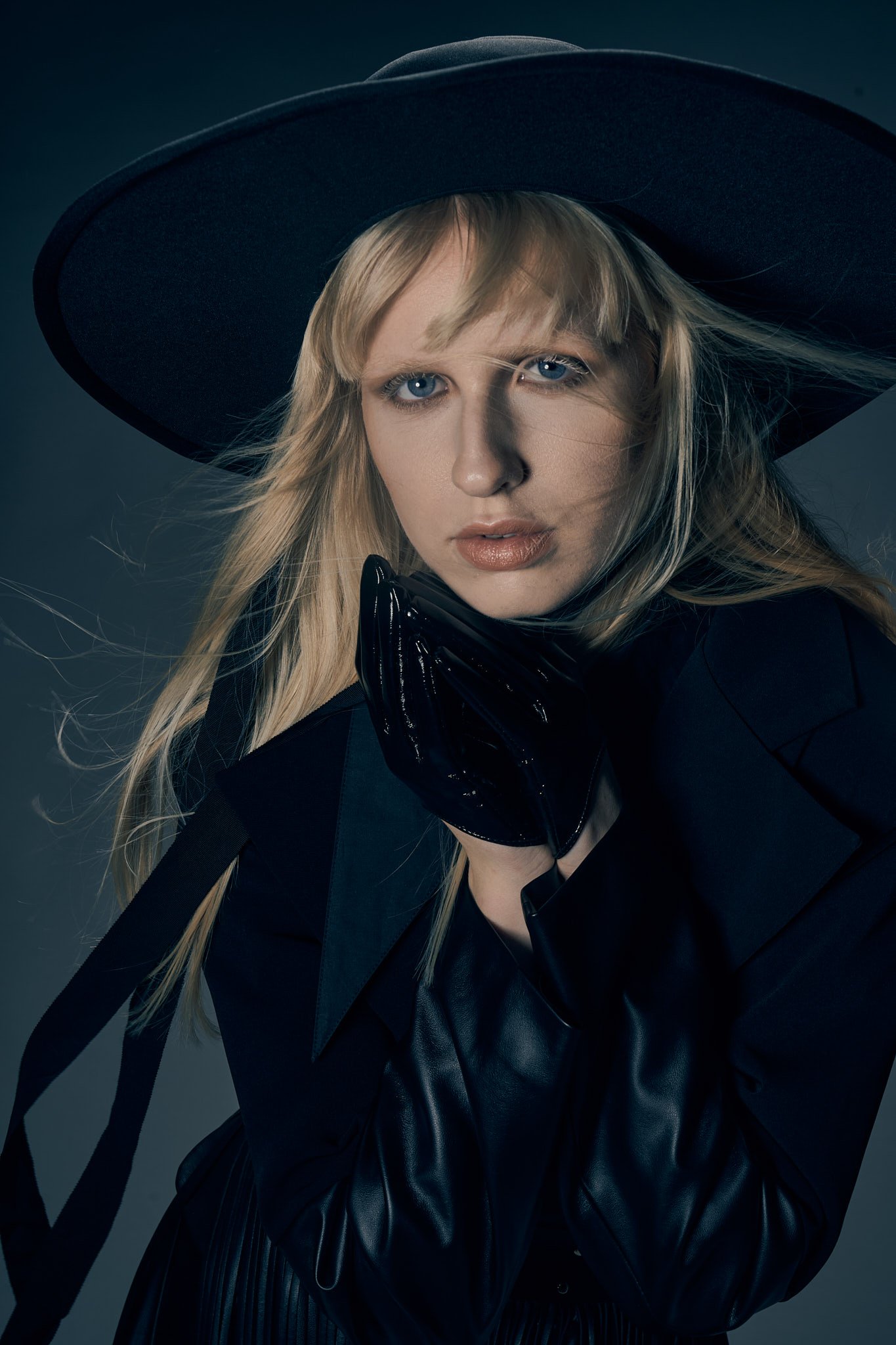 Close-up portrait of a blonde model wearing a wide-brim black felt hat, leather gloves, and a dark Western tailored coat.⁠