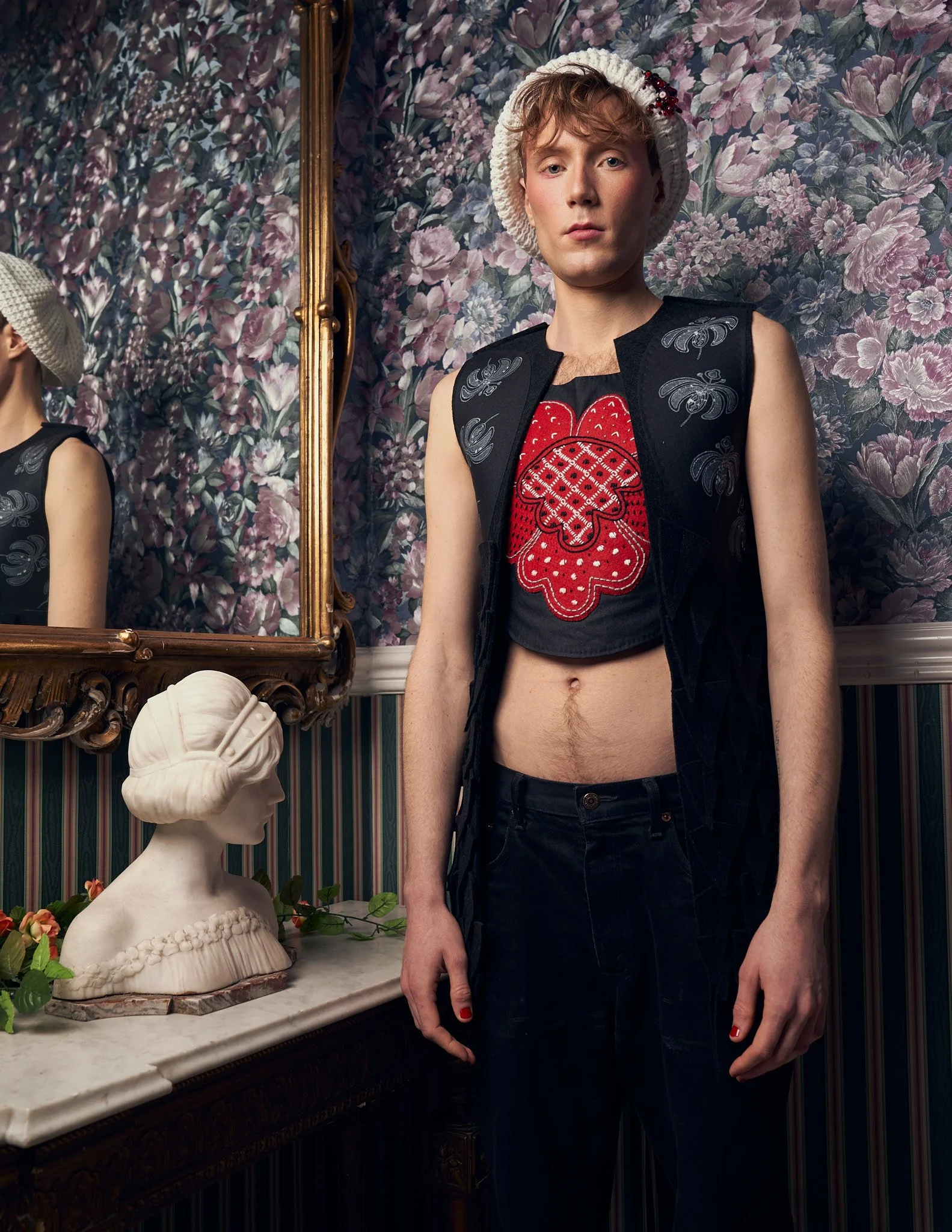 n editorial portrait of a male model in a white knit beanie, black open vest over a red embroidery-style cropped top, standing in an ornate hotel interior.⁠