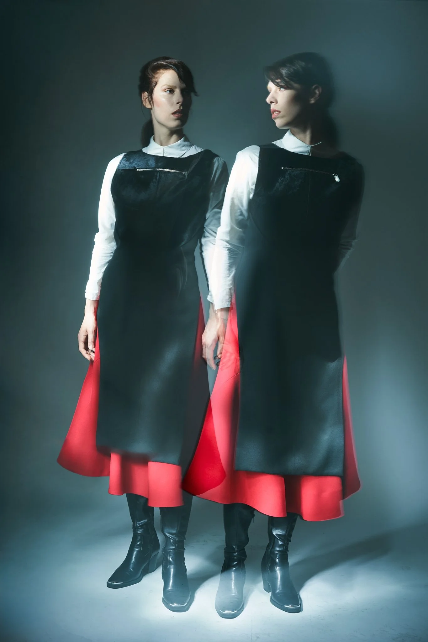 ⁠Fashion editorial multiple exposure studio photograph of a model standing side-by-side, wearing a structured black tunic over a long white shirt, and dynamic layered red and black skirt with black ankle boots.⁠