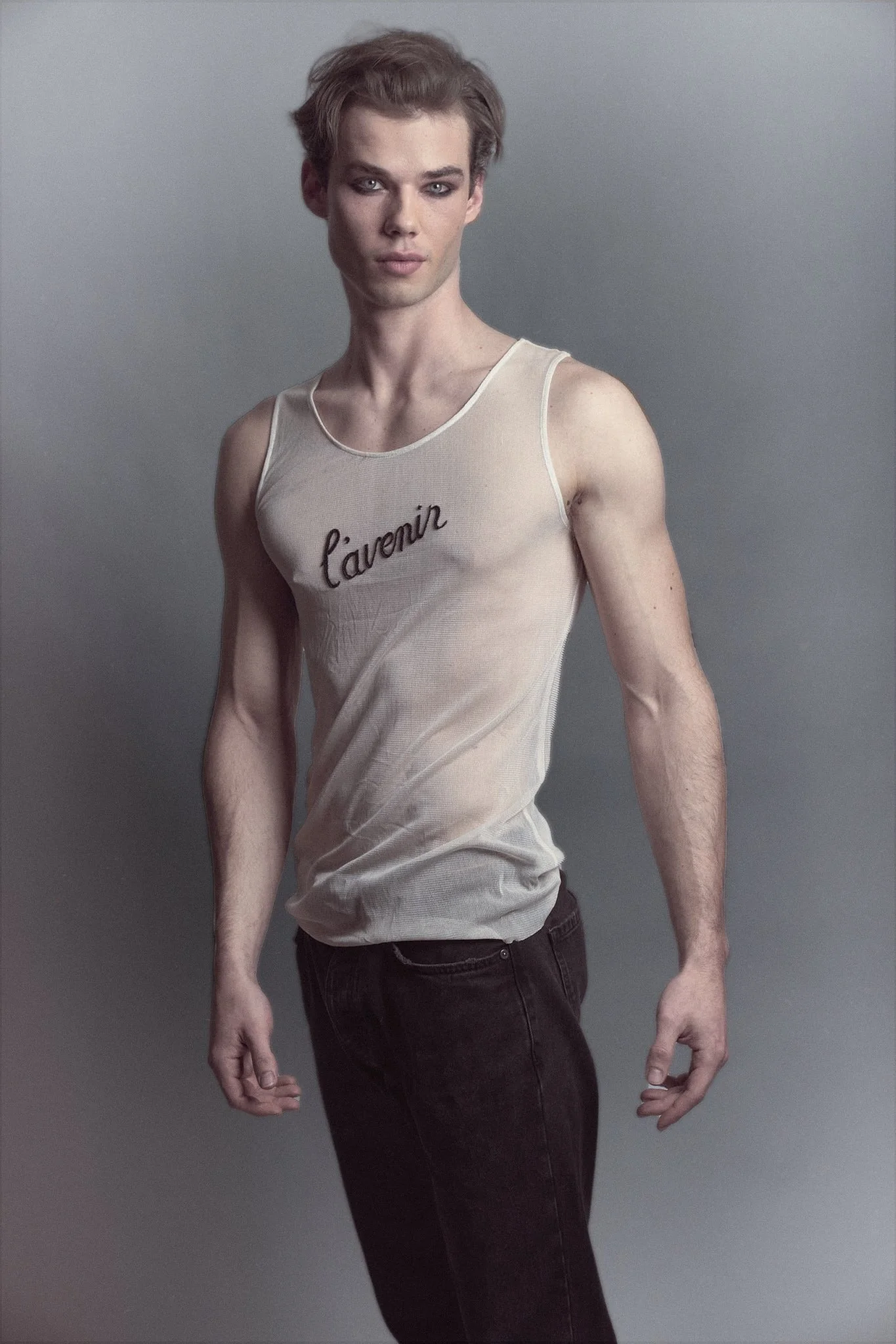Full-body studio portrait of a male model in a sheer white “l’avenir” Ann Demeulemeester tank top and black pants, standing in soft cinematic lighting.