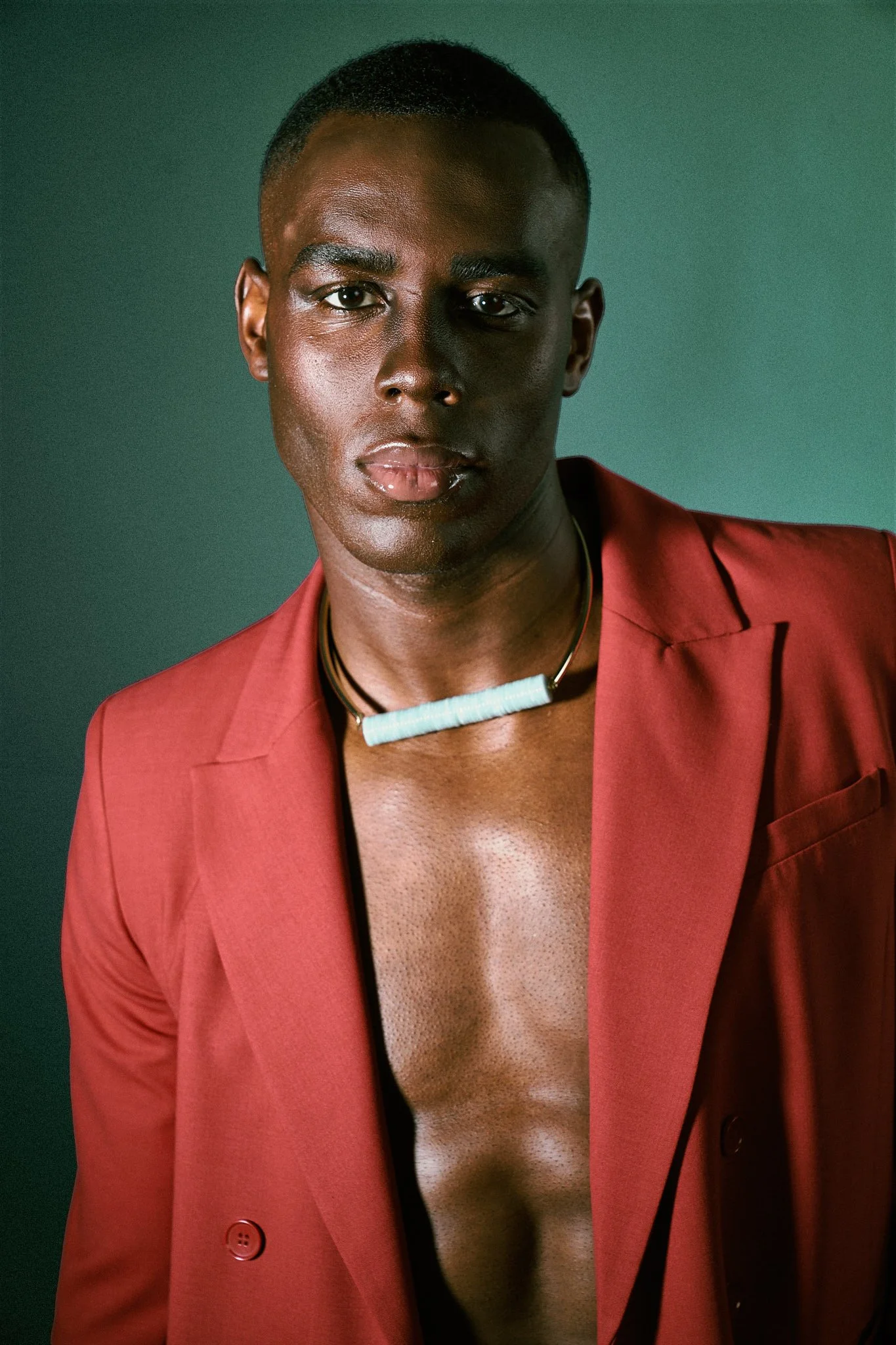 Close-up fashion portrait of a Black male model in a red blazer, shirtless, wearing a bold white block pendant necklace.⁠