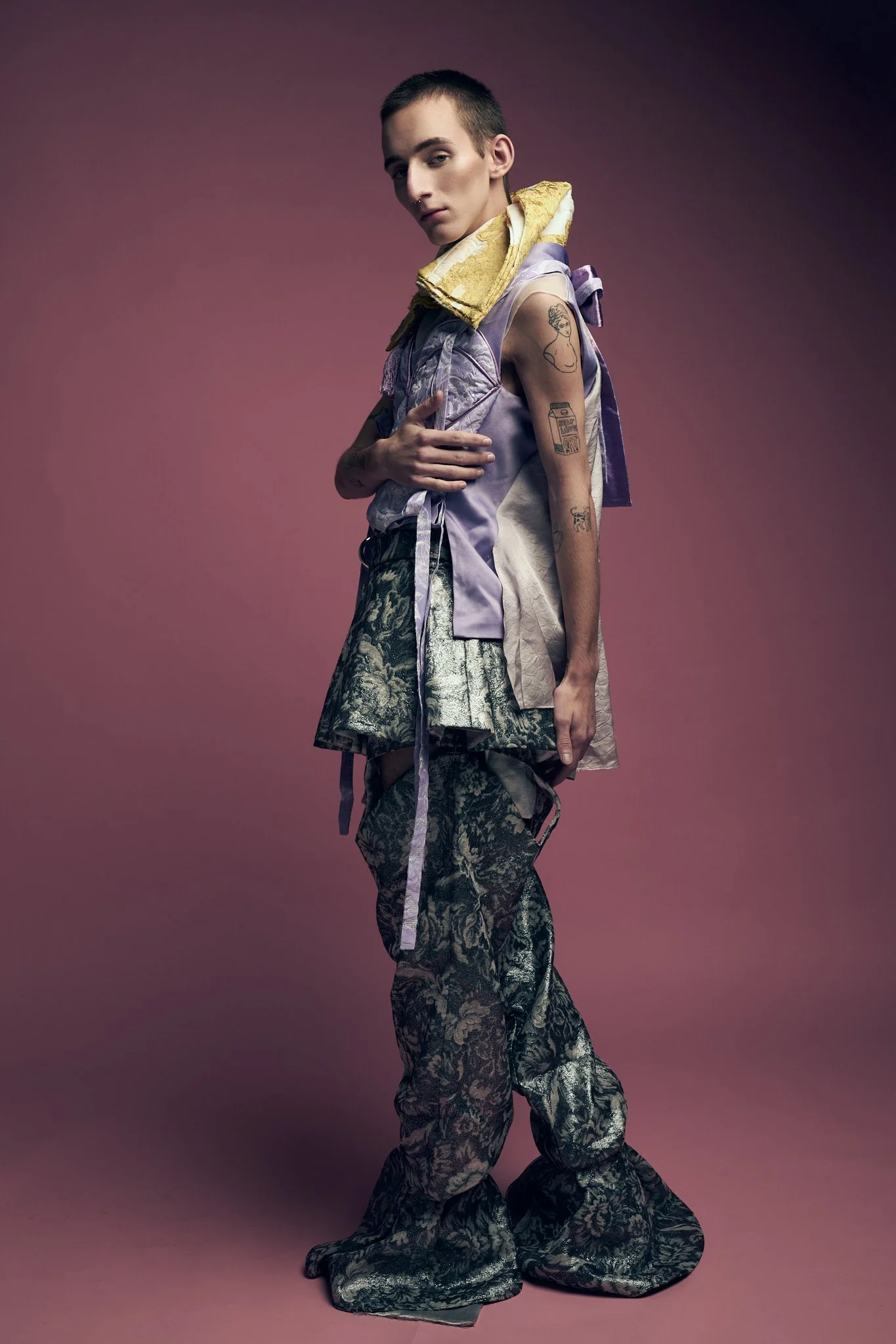 Full-length fashion portrait of a male model wearing a high-concept layered outfit with a yellow collar, lilac top, and textured dark flared trousers.⁠