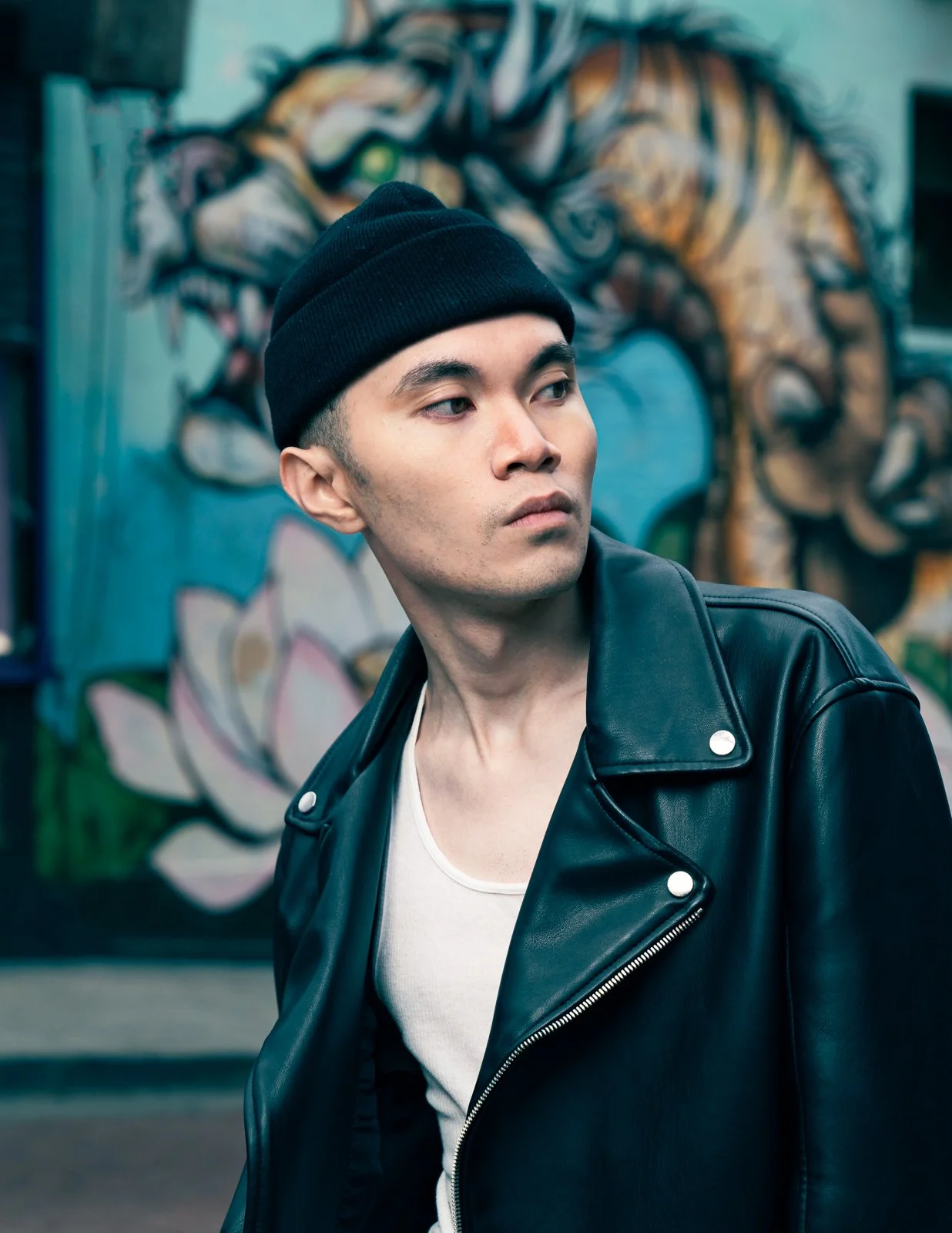 Editorial fashion portrait of a male model wearing a black beanie, white tank top, and black leather jacket in an urban setting by fashion photographer Simon Rogghe. 