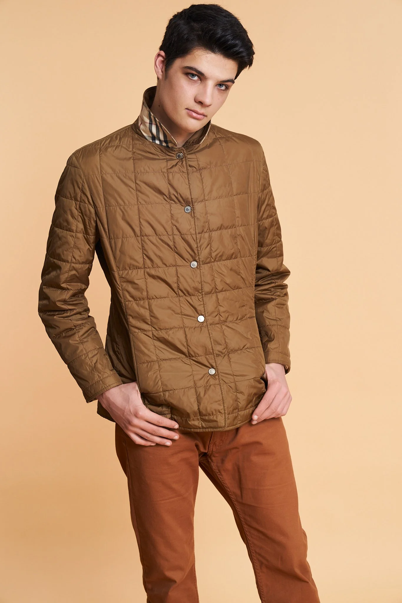Studio fashion portrait of a male model wearing a brown quilted jacket with tailored fit, photographed against a warm beige background, showcasing clean commercial styling and refined menswear photography.