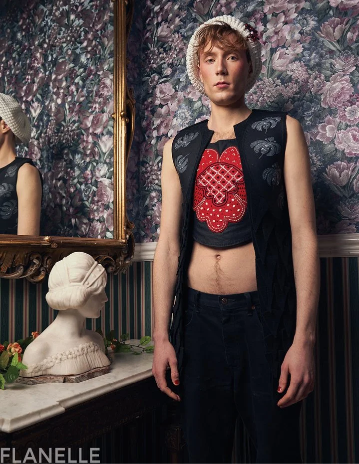Editorial menswear fashion portrait for Flanelle Magazine featuring a model in a crochet cap, embroidered vest, and dark trousers in a vintage floral interior.