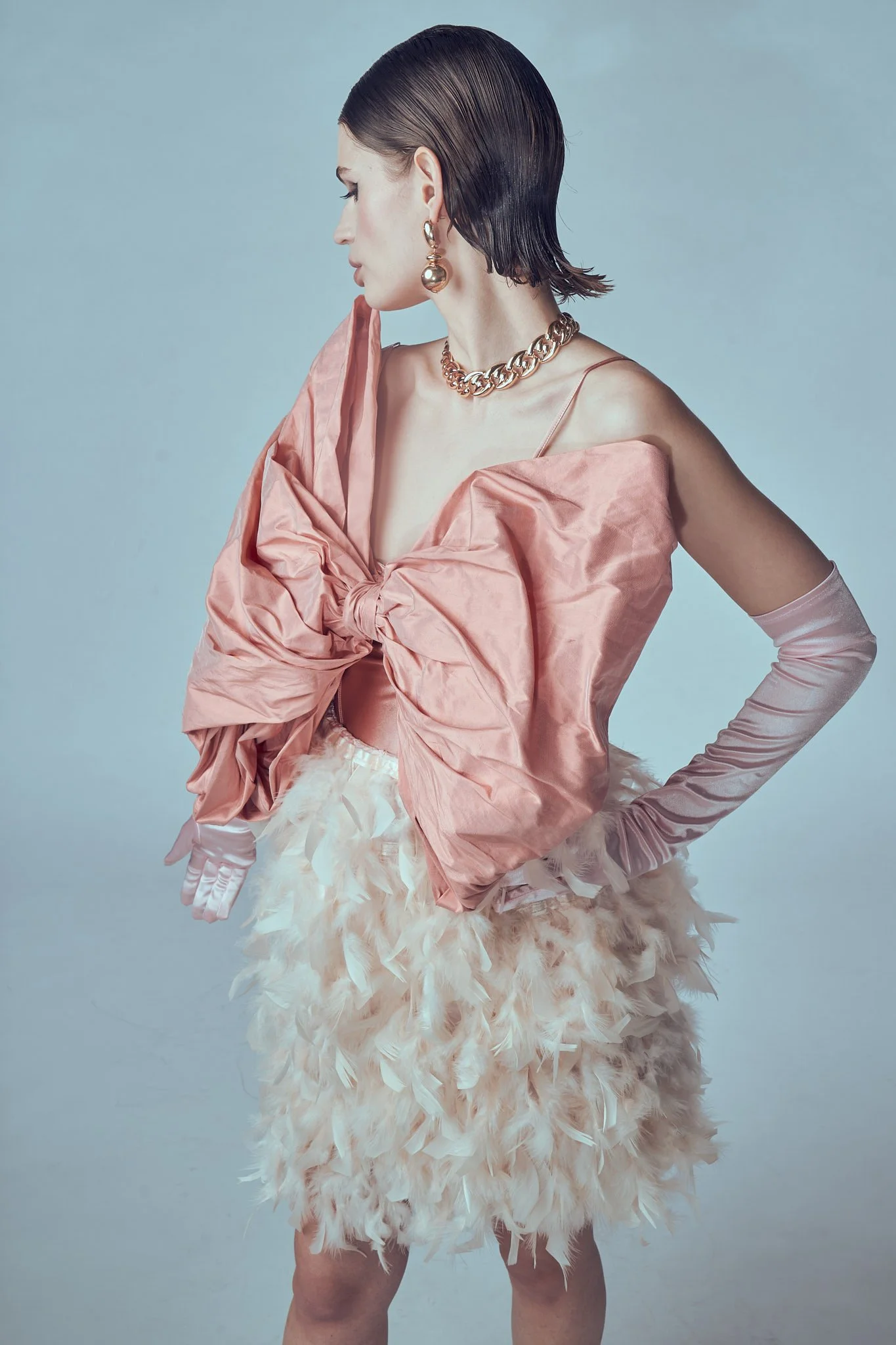 Three-quarter fashion portrait of a model looking away from the camera, wearing an oversized pastel pink bow top with matching opera gloves and a textured white feather mini skirt.