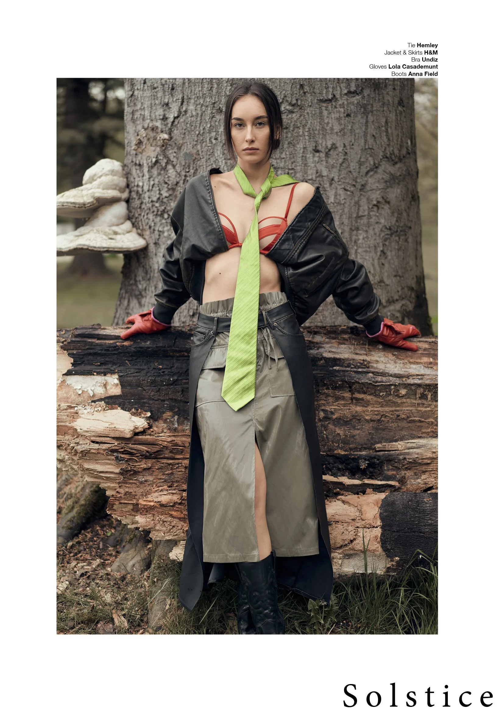 Editorial fashion photography of a female model leaning against a moss-covered tree trunk with large bracket fungi, wearing a black leather bomber jacket, olive green leather midi skirt, red leather gloves, and black cowboy boots, shot on location in