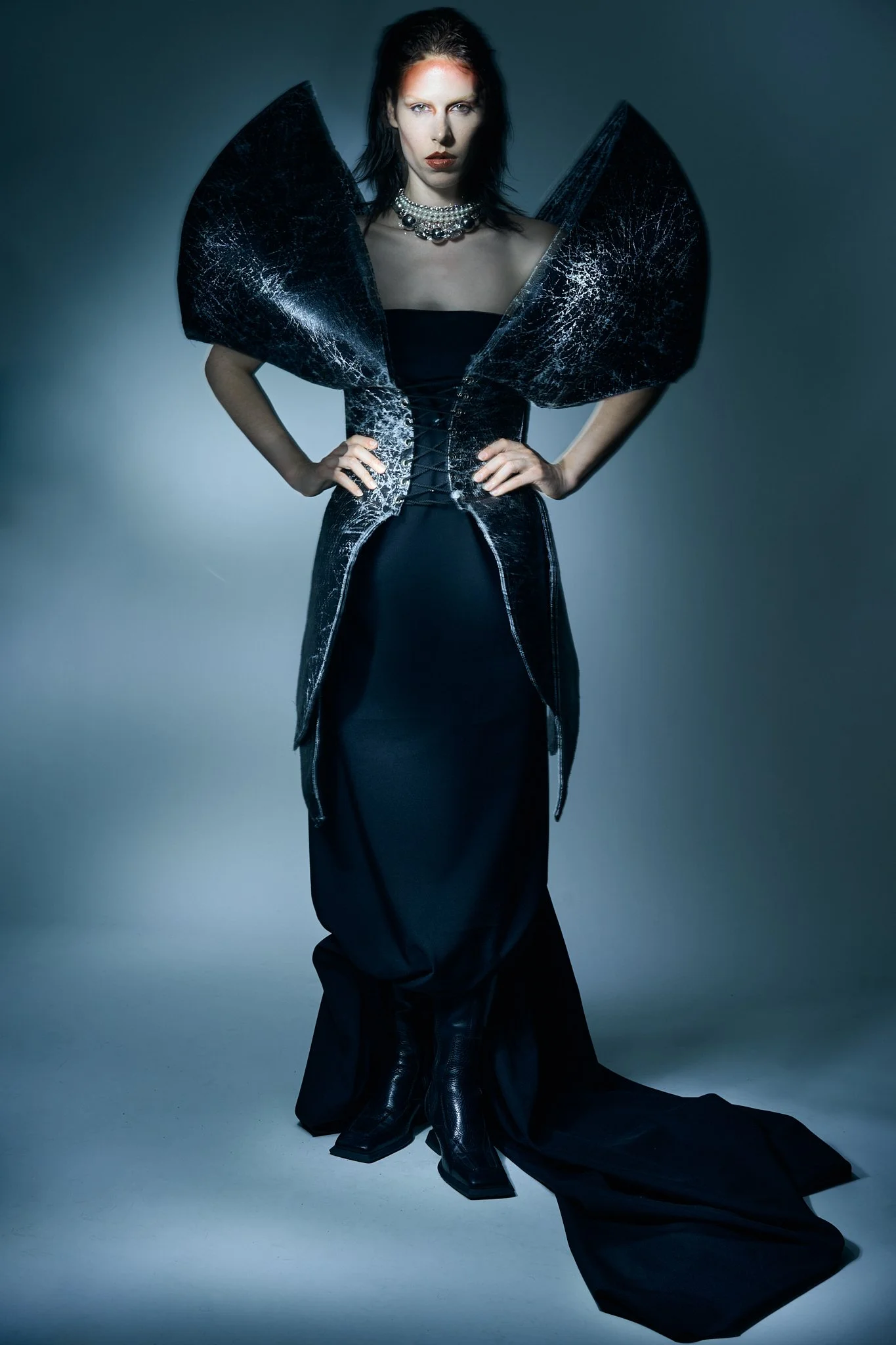 Full-length editorial photograph of a model in a dramatic structural gown with exaggerated shoulders and intricate texture.⁠