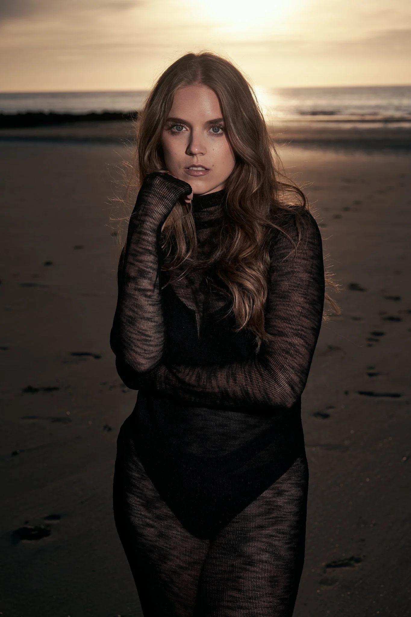 Woman in black sheer mesh bodysuit posing thoughtfully on beach with golden sunset behind, moody fashion photography by Ghent-based photographer Simon Rogghe