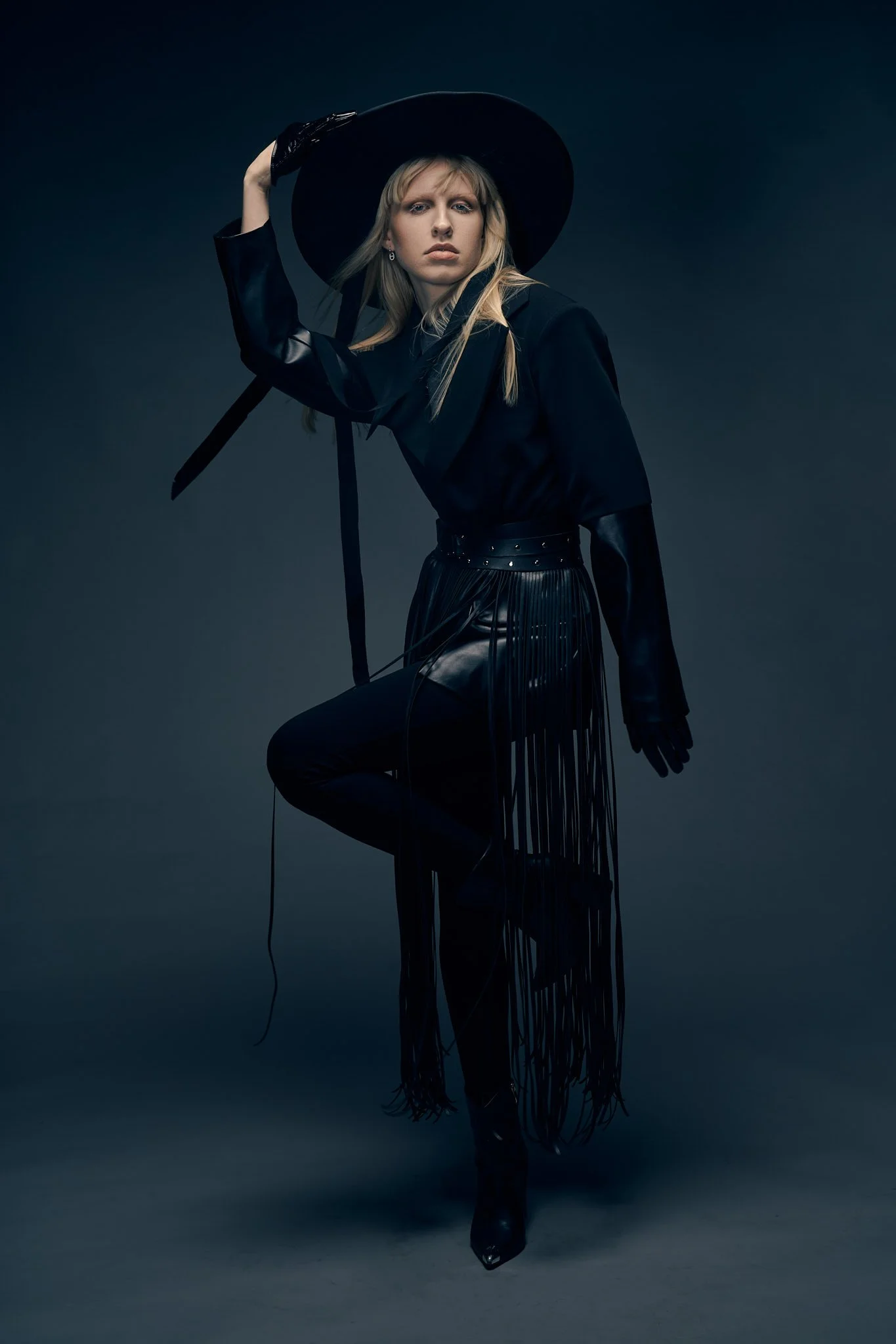 Full-length Western fashion editorial photo of a blonde model in a wide-brim black hat, dark tailored jacket, and black leather fringe trousers.⁠