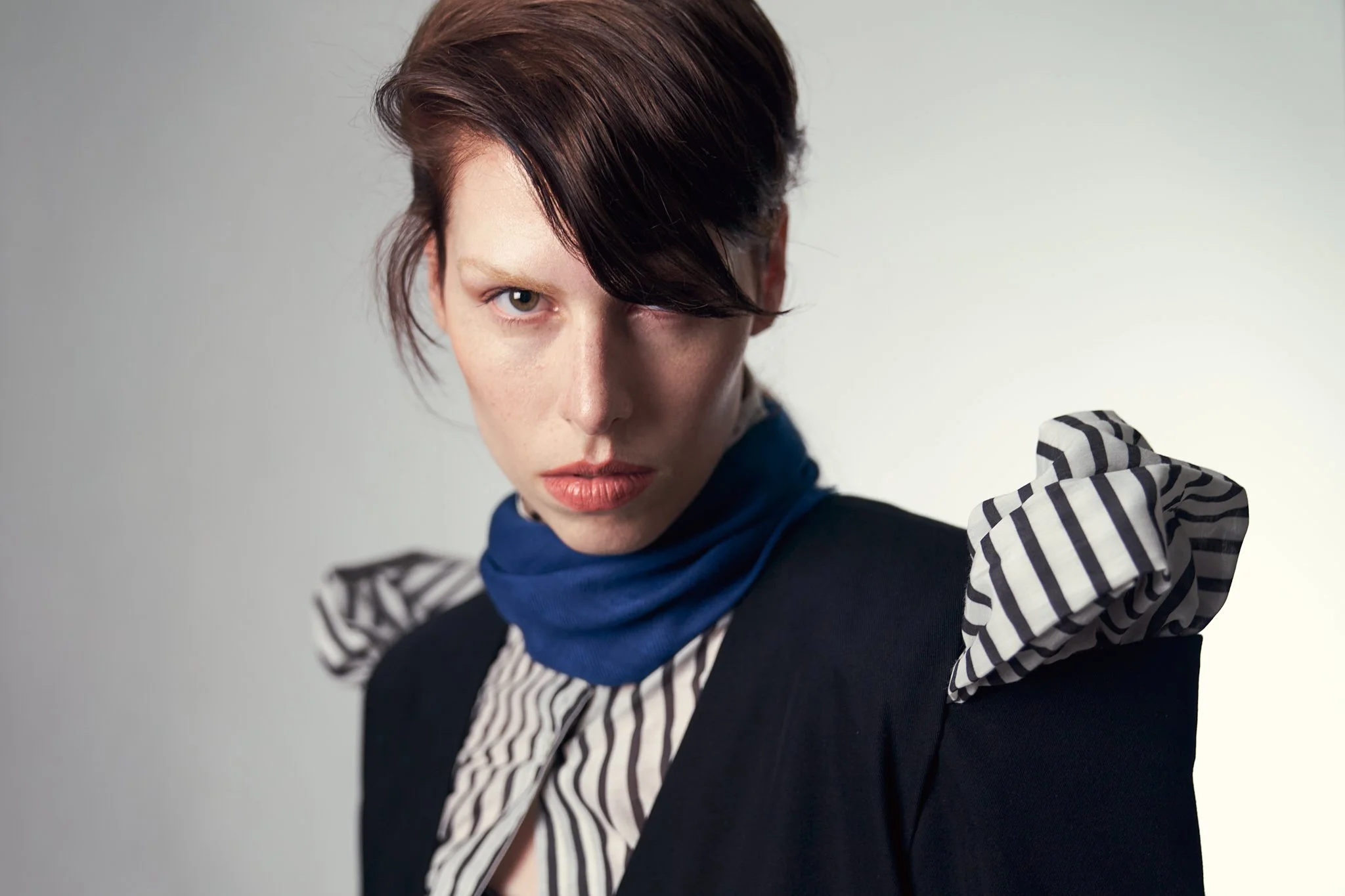 Fashion editorial close-up portrait of a model with short dark hair wearing a structural black blazer with distinctive white and black striped ruffles on the shoulder.⁠