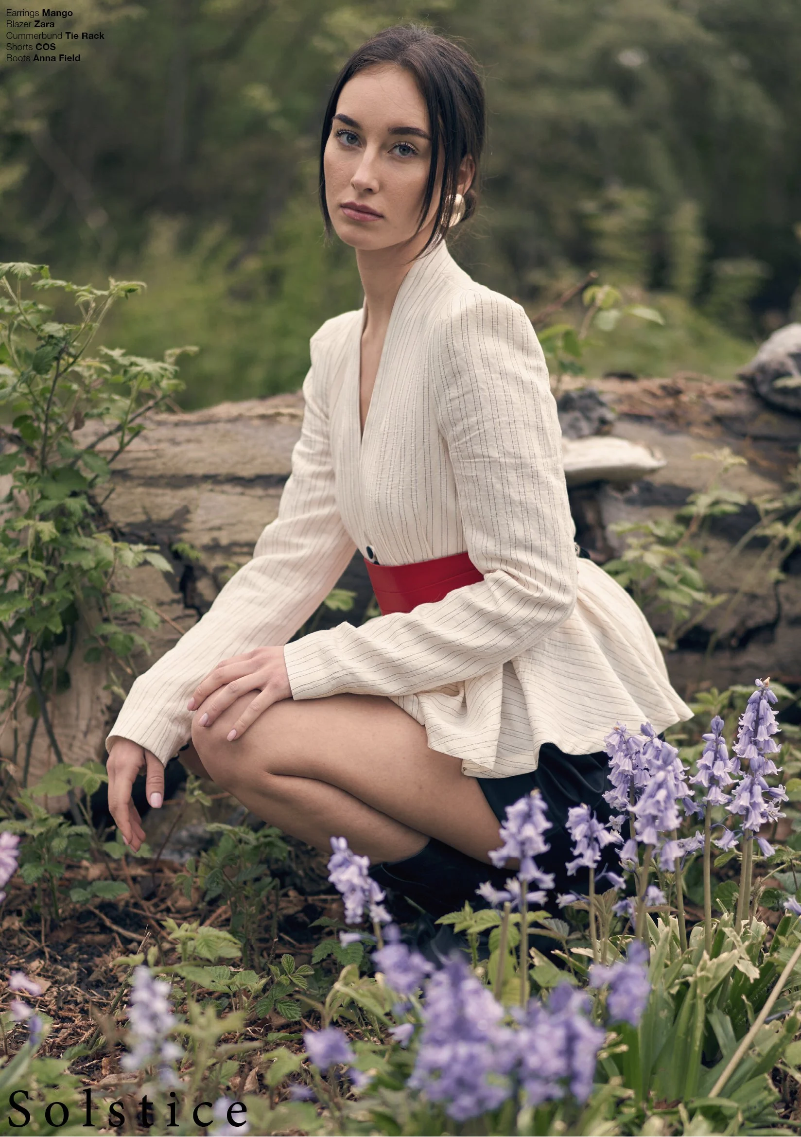 Elegant fashion portrait of a model seated by a woodland log wearing a structured white mini dress with deep neckline, bold red waist belt, and black knee-high boots. Sophisticated outdoor styling blends modern tailoring with natural surroundings in 